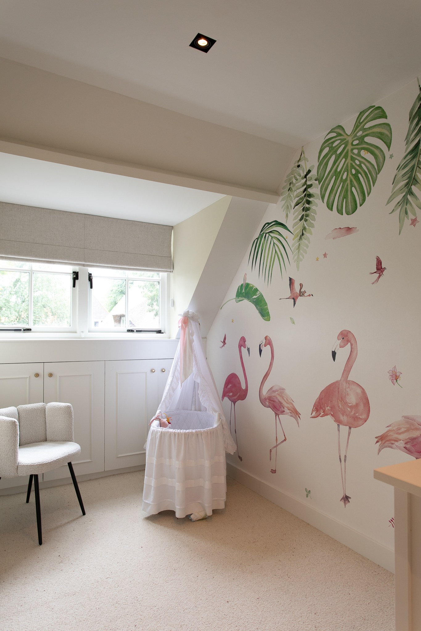 Lichte babykamer met speels flamingo- en palmbladbehang in roze en groen, witte lambrisering, naturel wieg en zacht crèmekleurig tapijt.