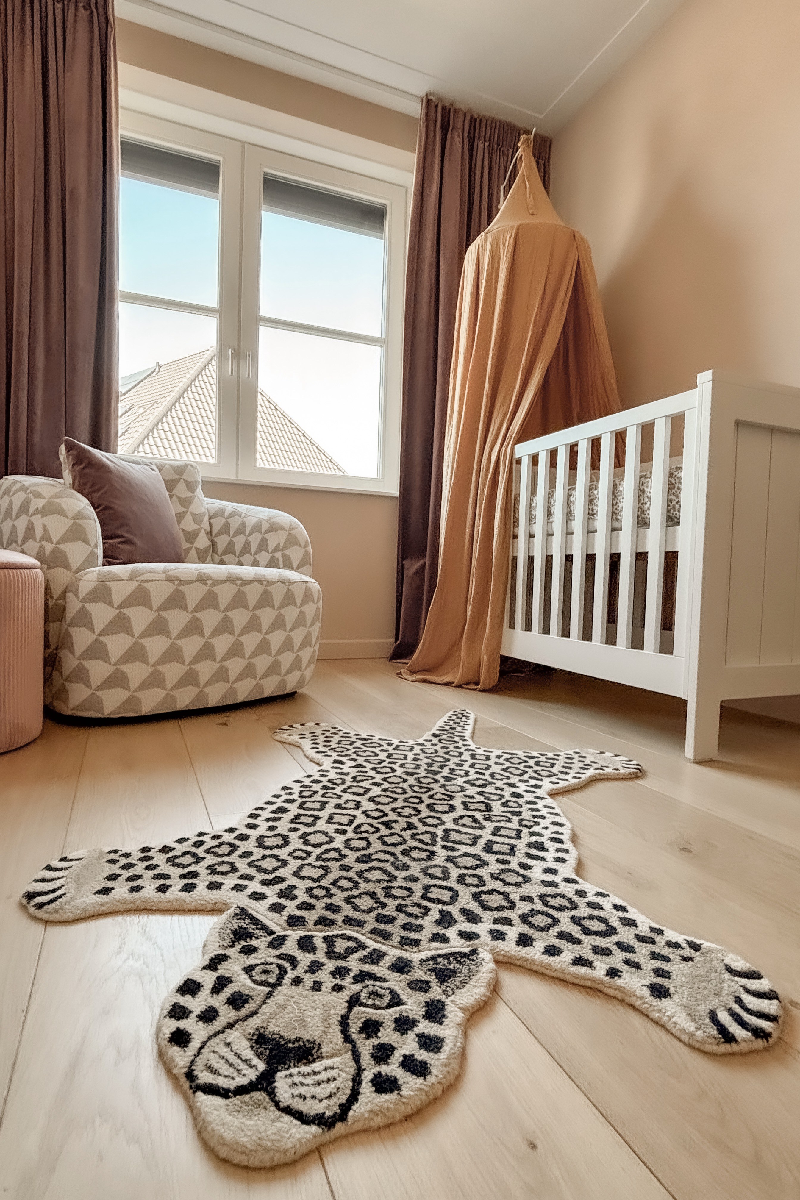 Warme babykamer met wit ledikant, bruine gordijnen, luipaardvloerkleed en roze bedhemel.