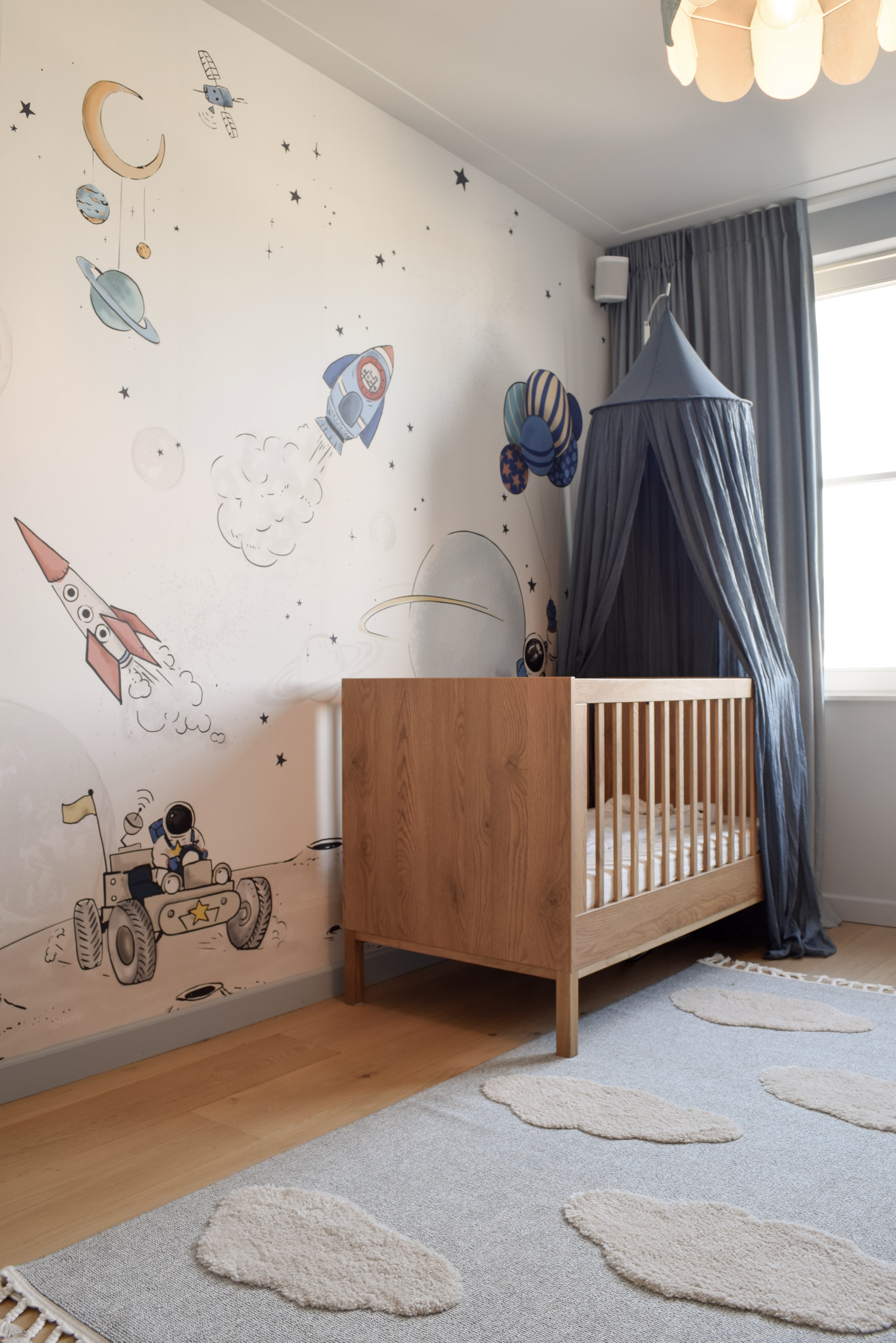Babykamer met ruimtethema op behang, eiken ledikant en donkerblauwe bedhemel.