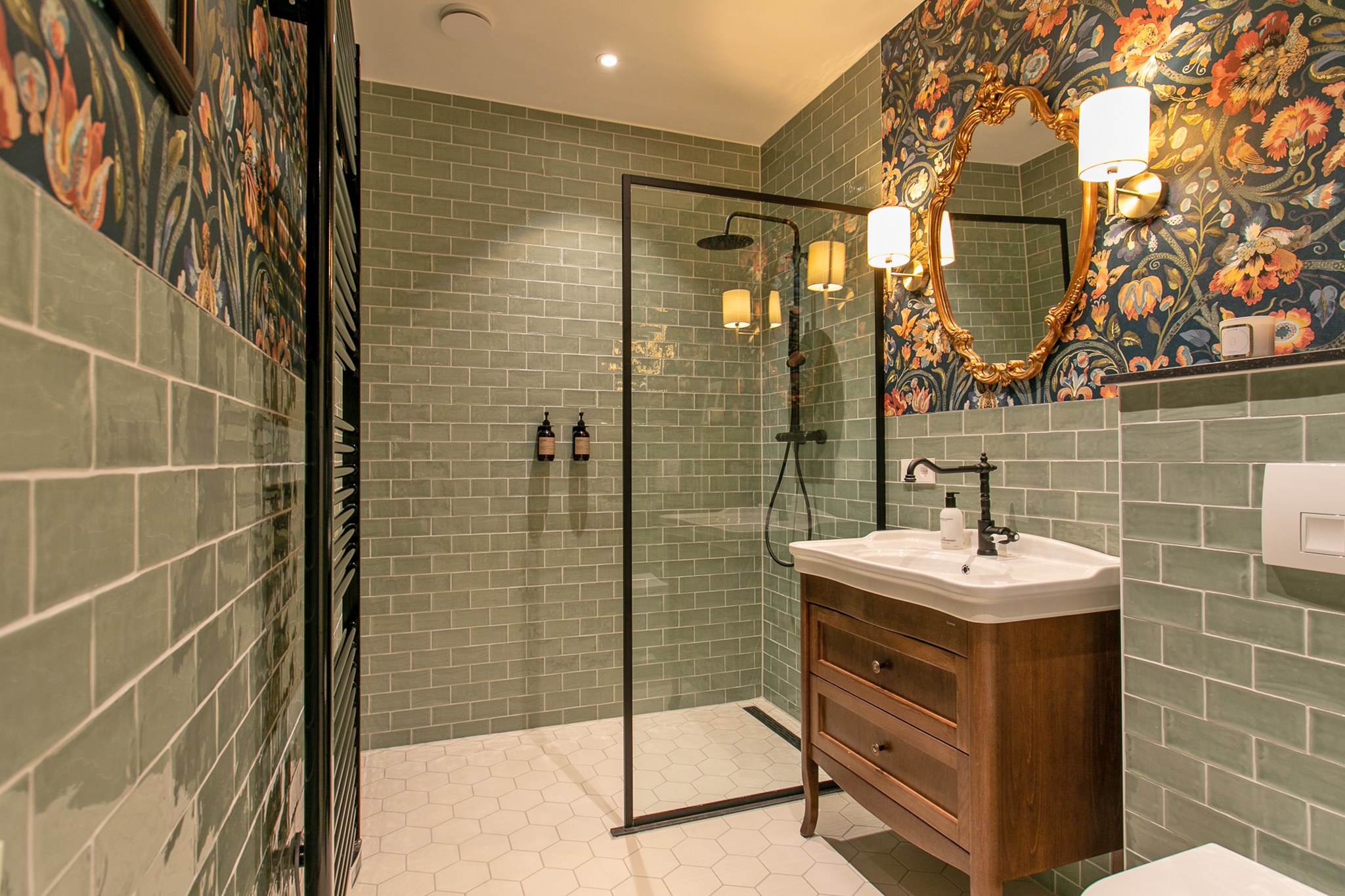 Sfeervolle badkamer met grijsgroene metrotegels, botanisch donker behang, walnoot wastafelmeubel, zwarte douchewand en goudkleurige spiegel en wandlampen.