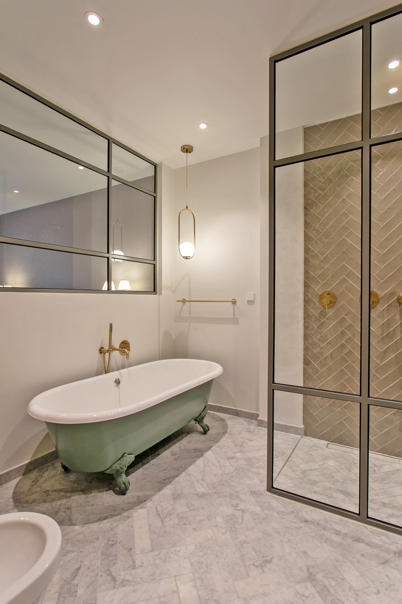Luxe badkamer met zachtgroen vrijstaand bad op pootjes, marmerlook vloer, zwarte stalen douchewand, messing kranen en wand met visgraattegels.