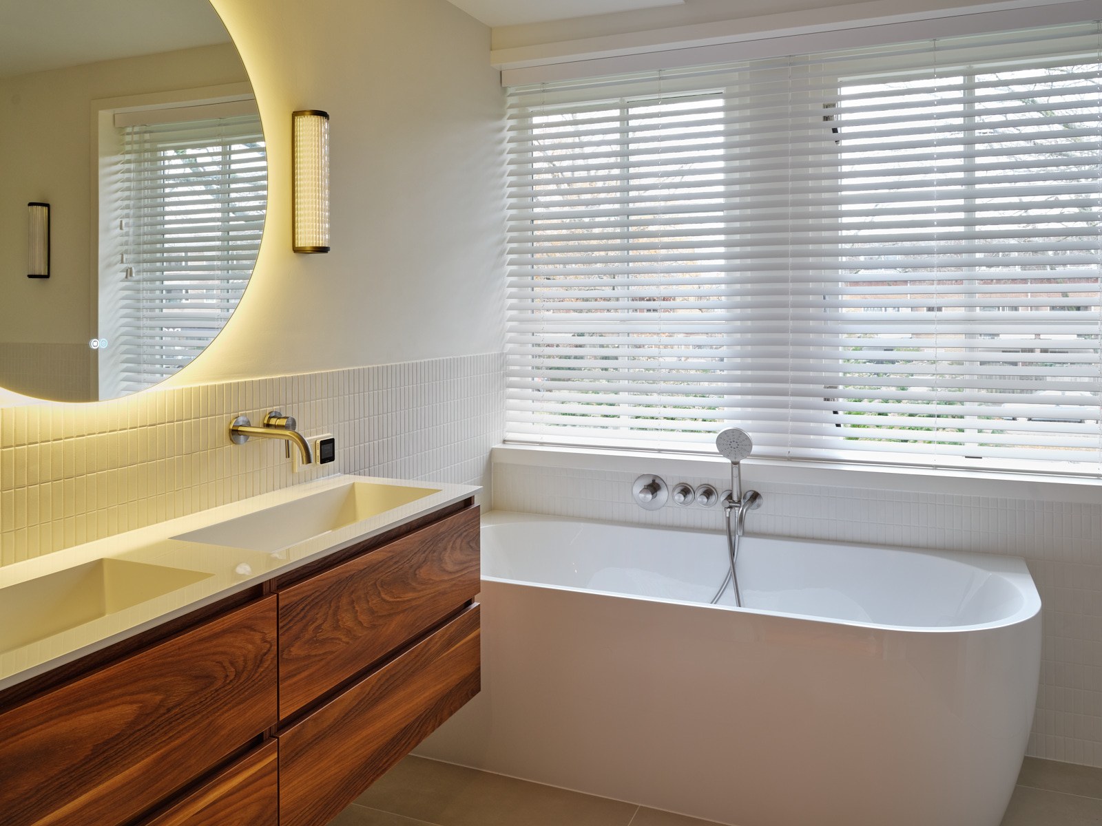 Lichte badkamer in Bussum met vrijstaand wit bad, zwevend walnoot wastafelmeubel, ronde spiegel met indirect licht, witte tegels en messing kranen.