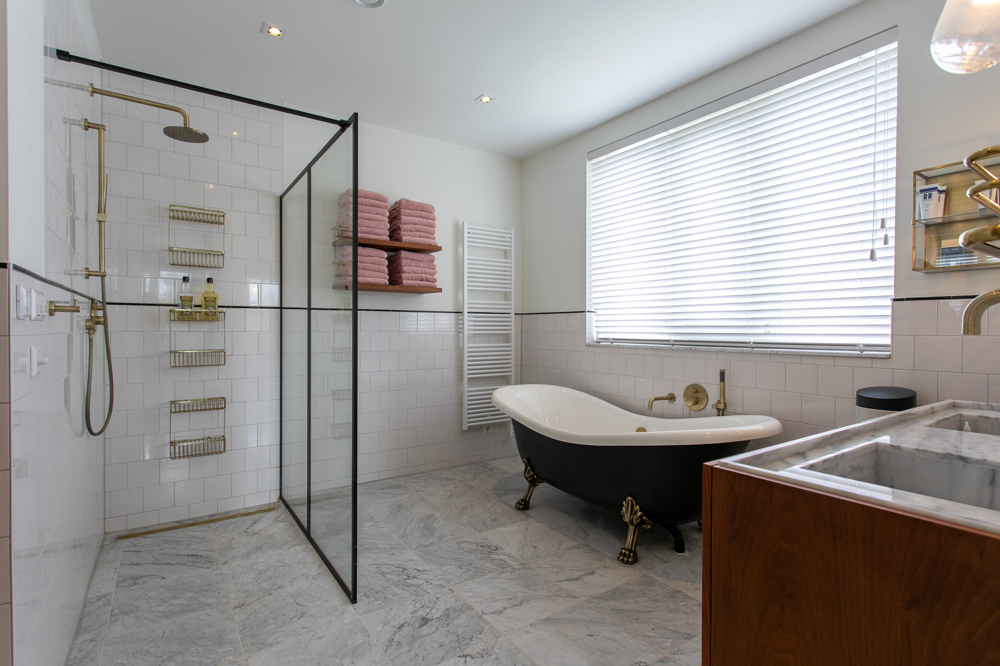 Luxe badkamer met zwart vrijstaand bad op messing pootjes, inloopdouche met zwarte omlijsting, marmerlook vloer, witte tegels en walnoot wastafelmeubel.