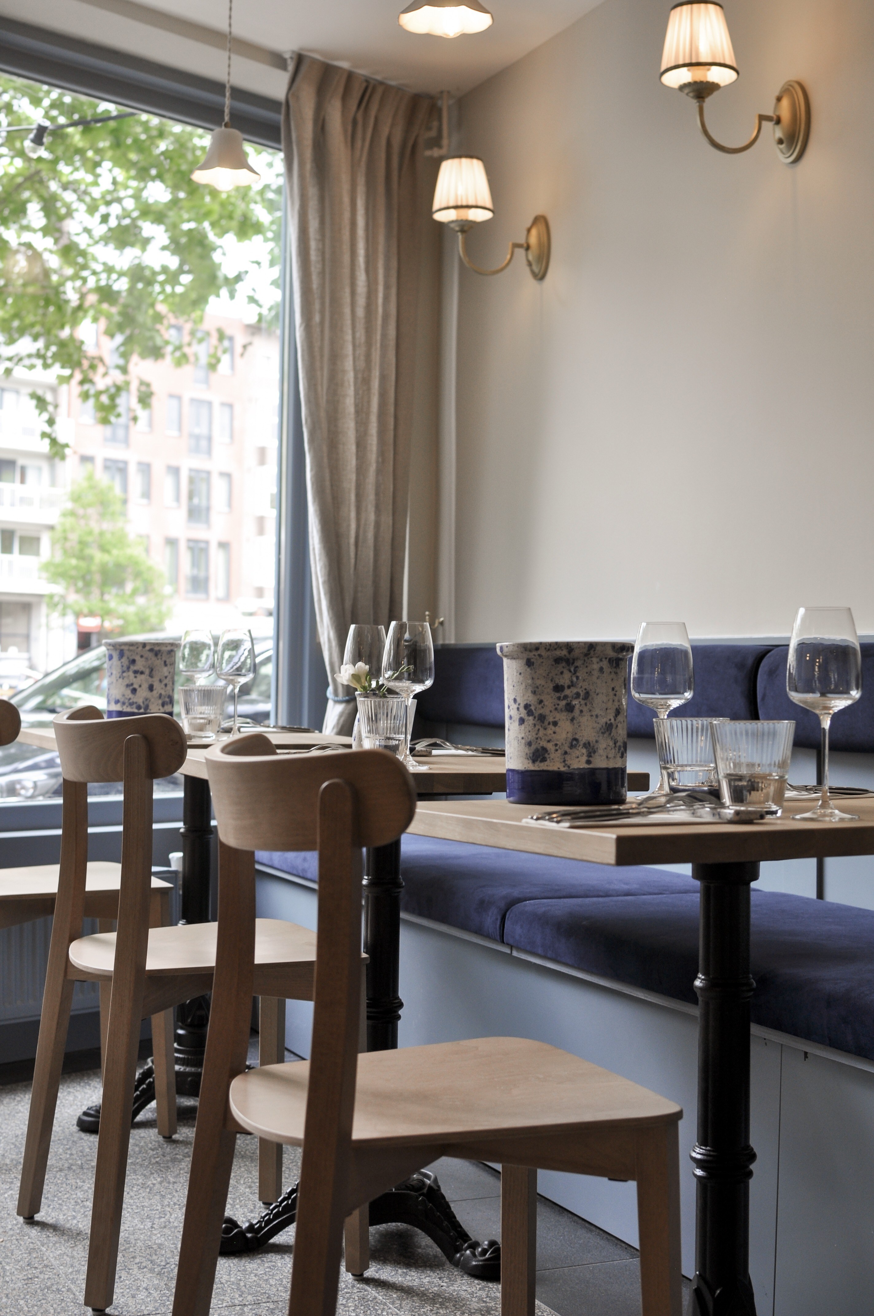 Caféhoek met donkerblauwe wandbank, houten stoelen, ronde tafels en klassieke wandlampen.