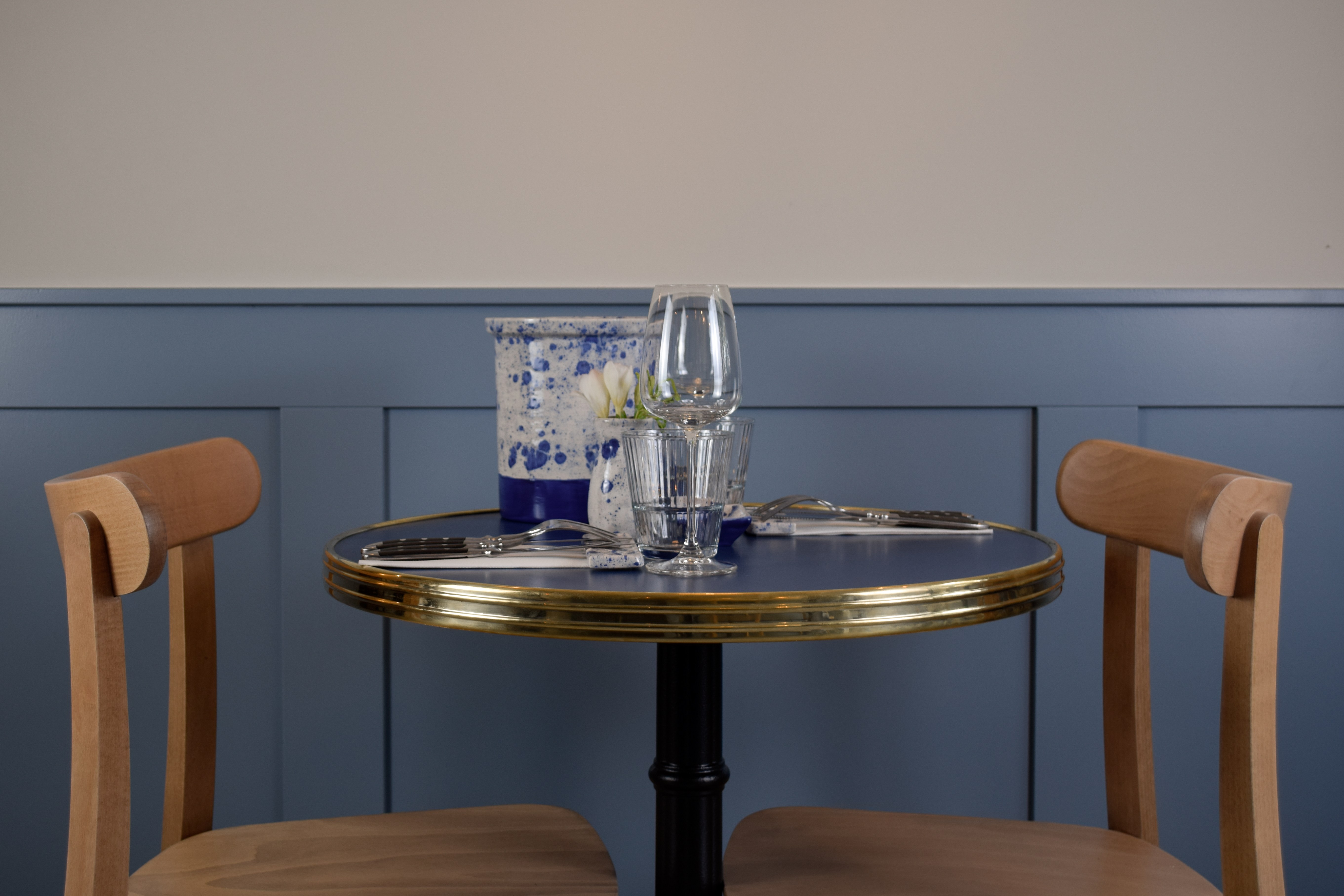 Tafeldetail in café met blauwe gepaneelde zitbank, messing tafelrand en houten stoelen.