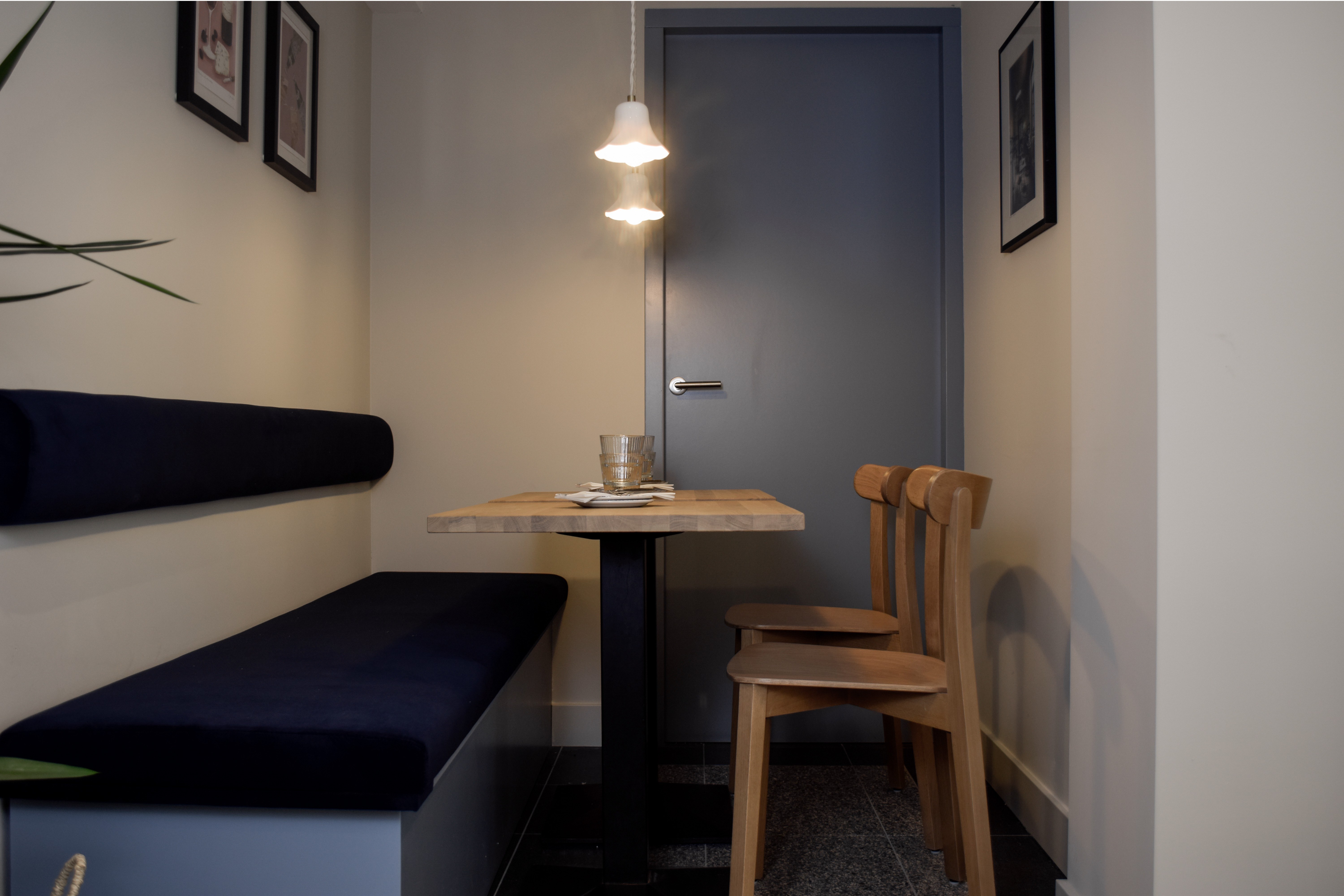 Intieme nis met donkerblauwe zitbank, houten tafel en stoelen, grijsblauwe deur en hanglampen.