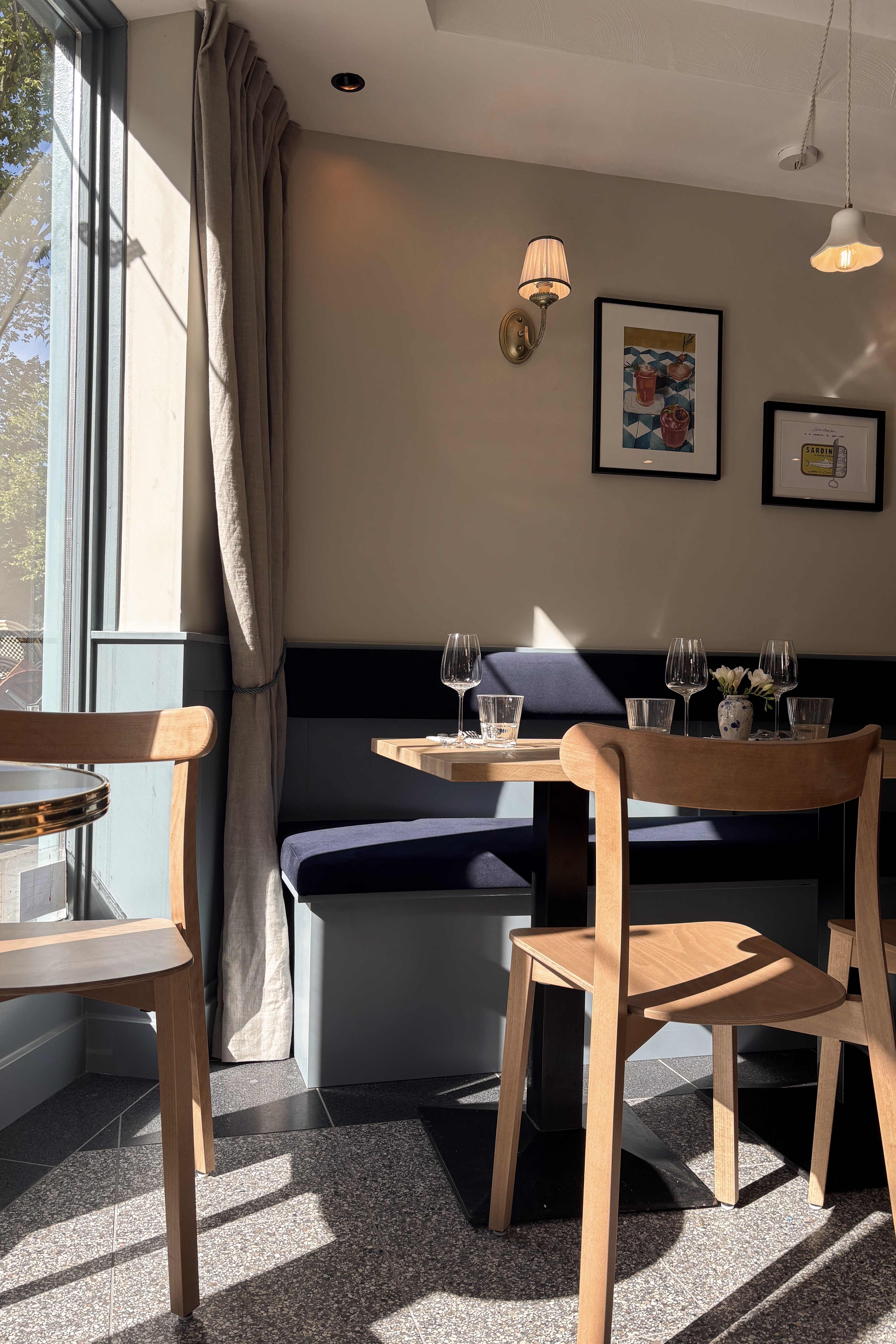Zonnige caféhoek met donkerblauwe wandbank, houten stoelen, ronde tafel en grote ramen.