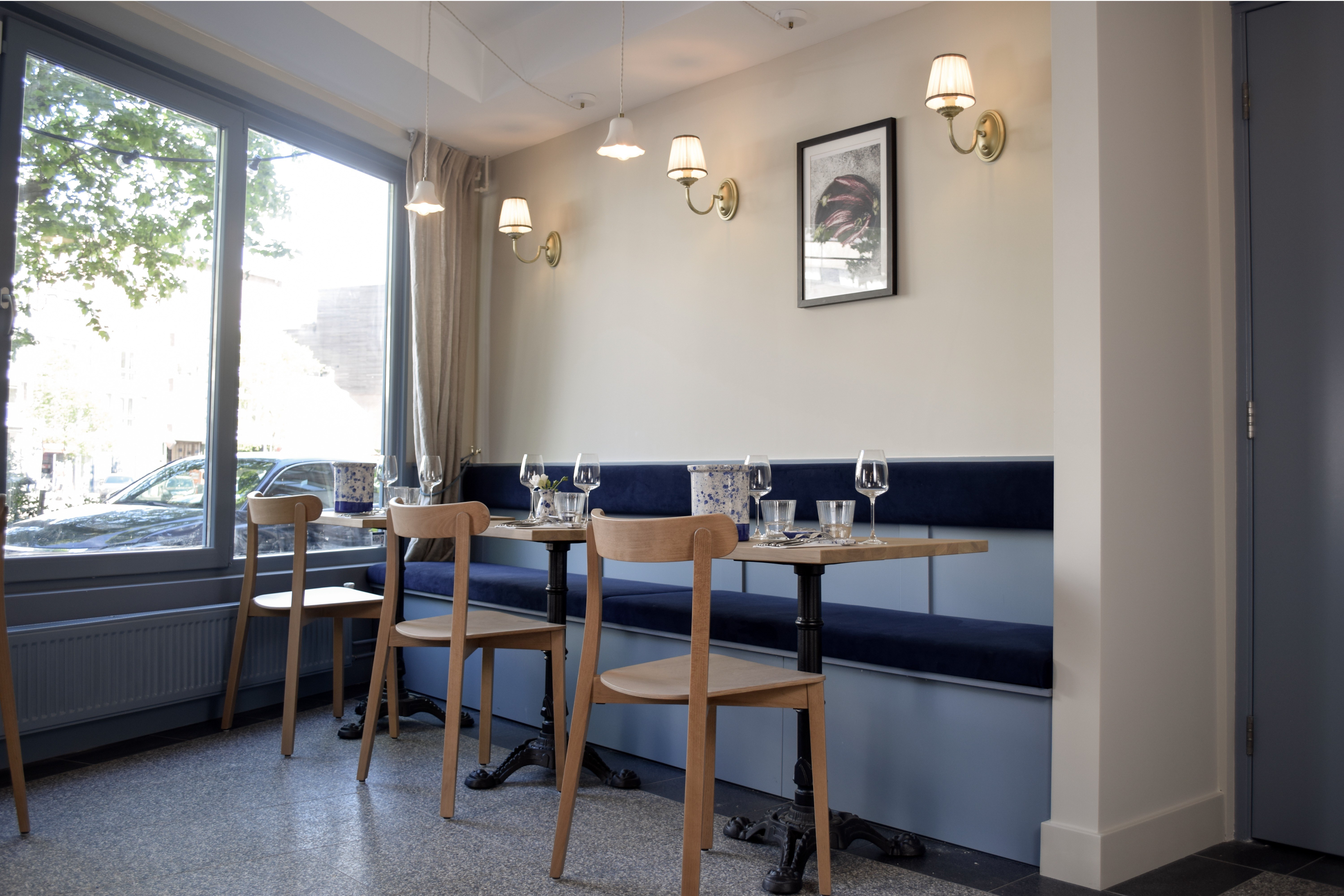 Lange donkerblauwe gepaneelde wandbank met ronde tafels, houten stoelen en grote ramen.