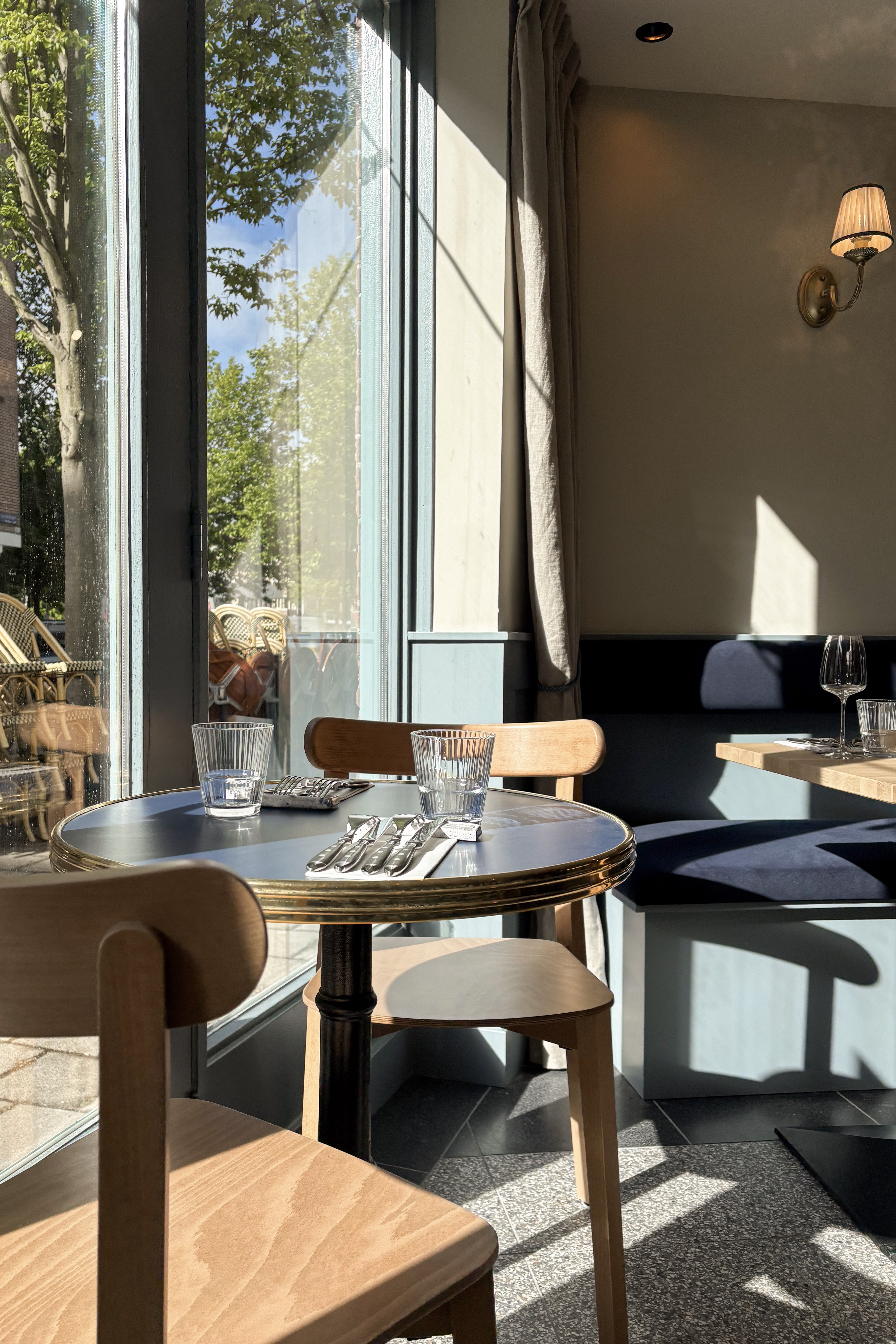 Zonnige caféhoek met ronde tafel met messing rand, houten stoelen, blauwe wandbank en grote ramen.