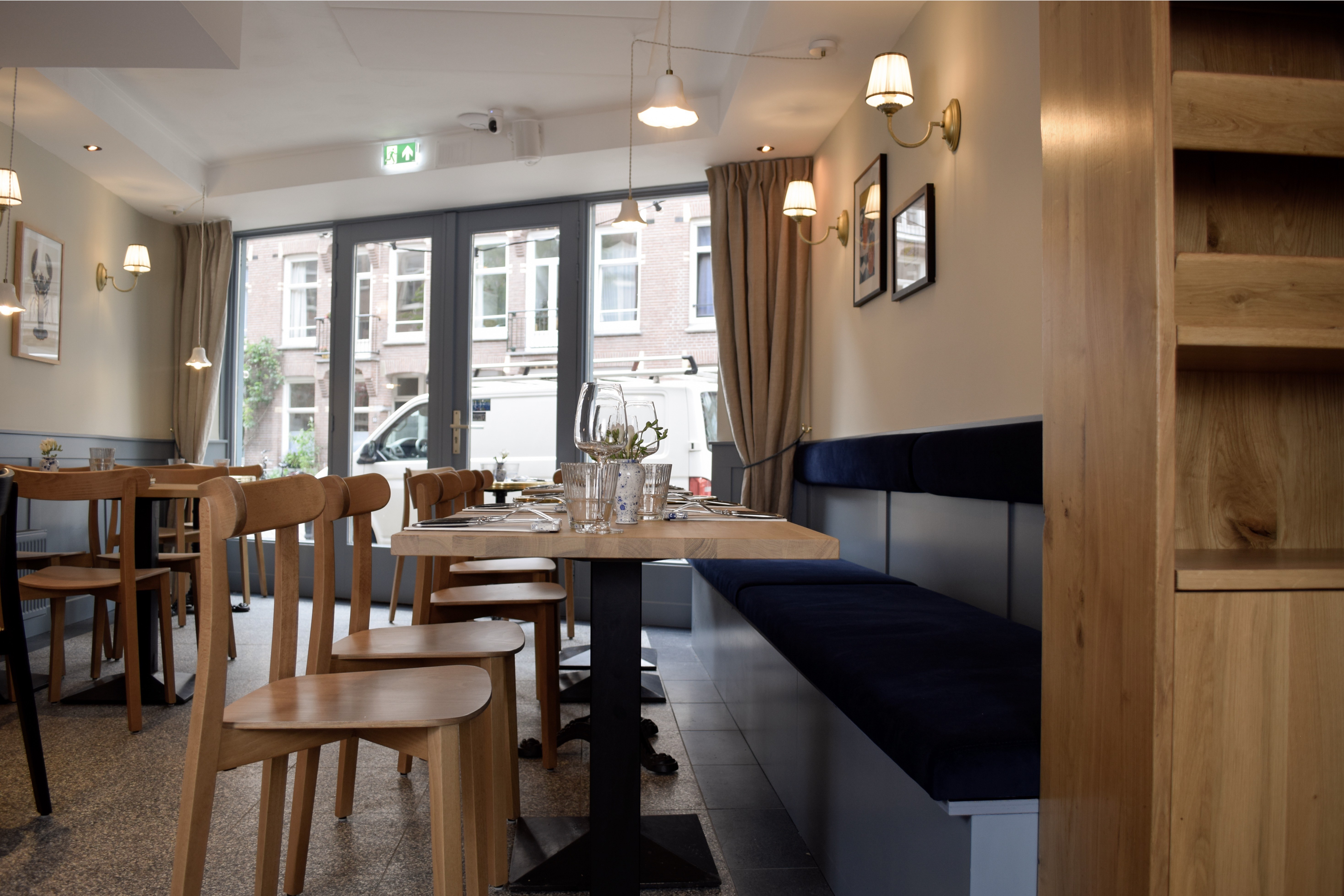 Interieur met lange donkerblauwe wandbank, houten tafels en stoelen, natuurlijke houtkast en grote voorramen.