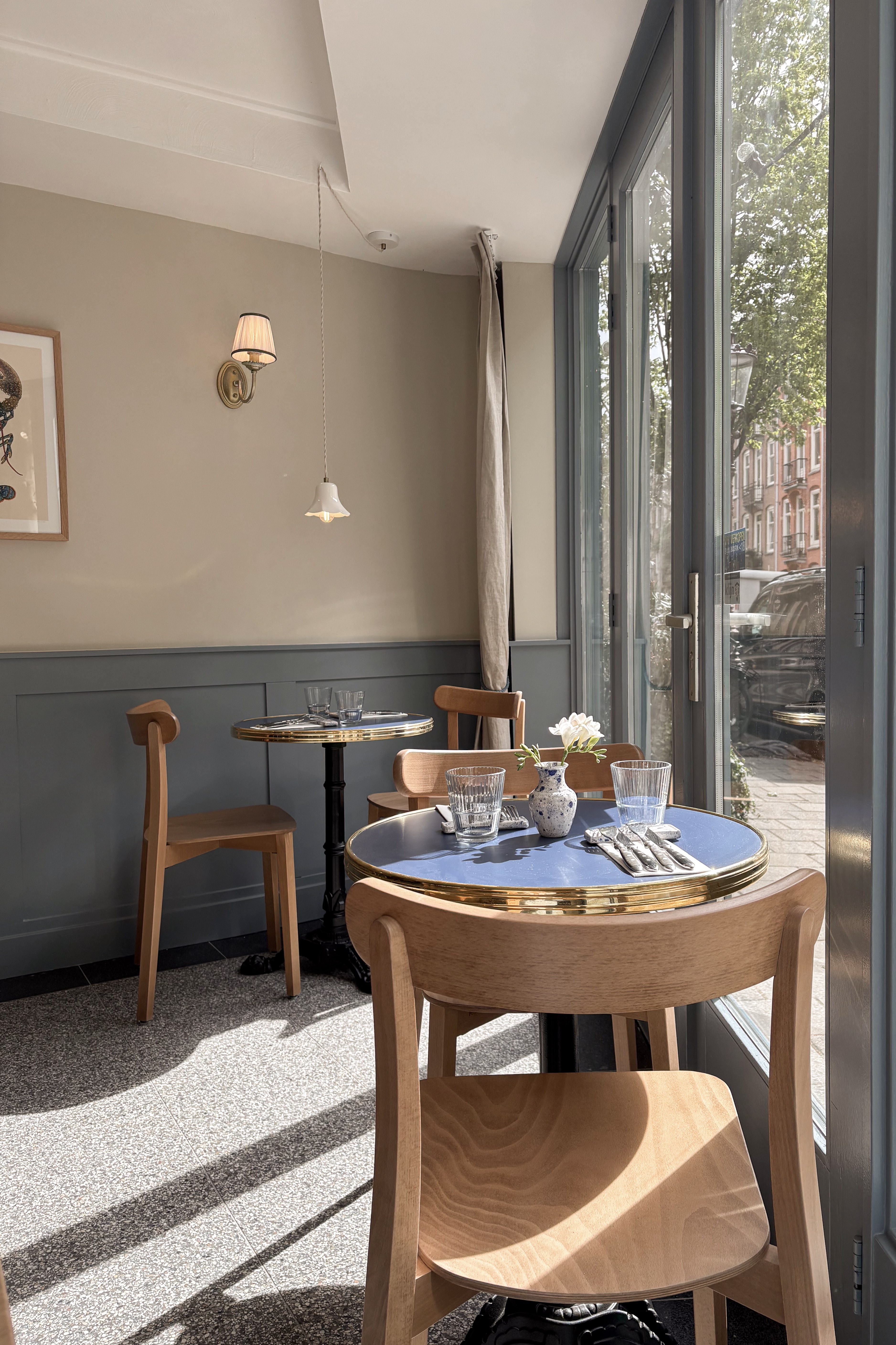 Lichte caféhoek met houten stoelen, ronde tafels, blauwgrijze lambrisering en kamerhoge glazen pui.