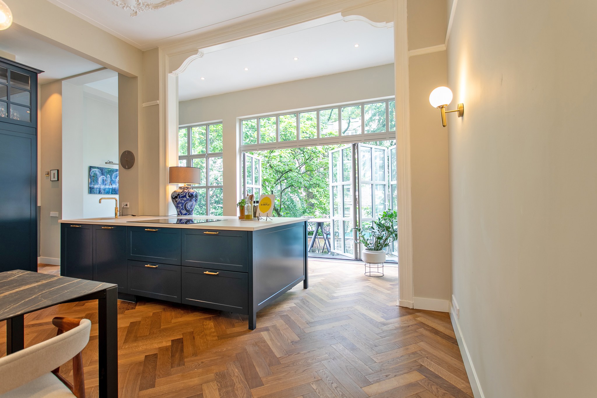 Ruime keuken met donkerblauw maatwerk kookeiland en kastenwand, messing grepen, licht natuurstenen werkblad, visgraatvloer en hoge glazen achterpui.