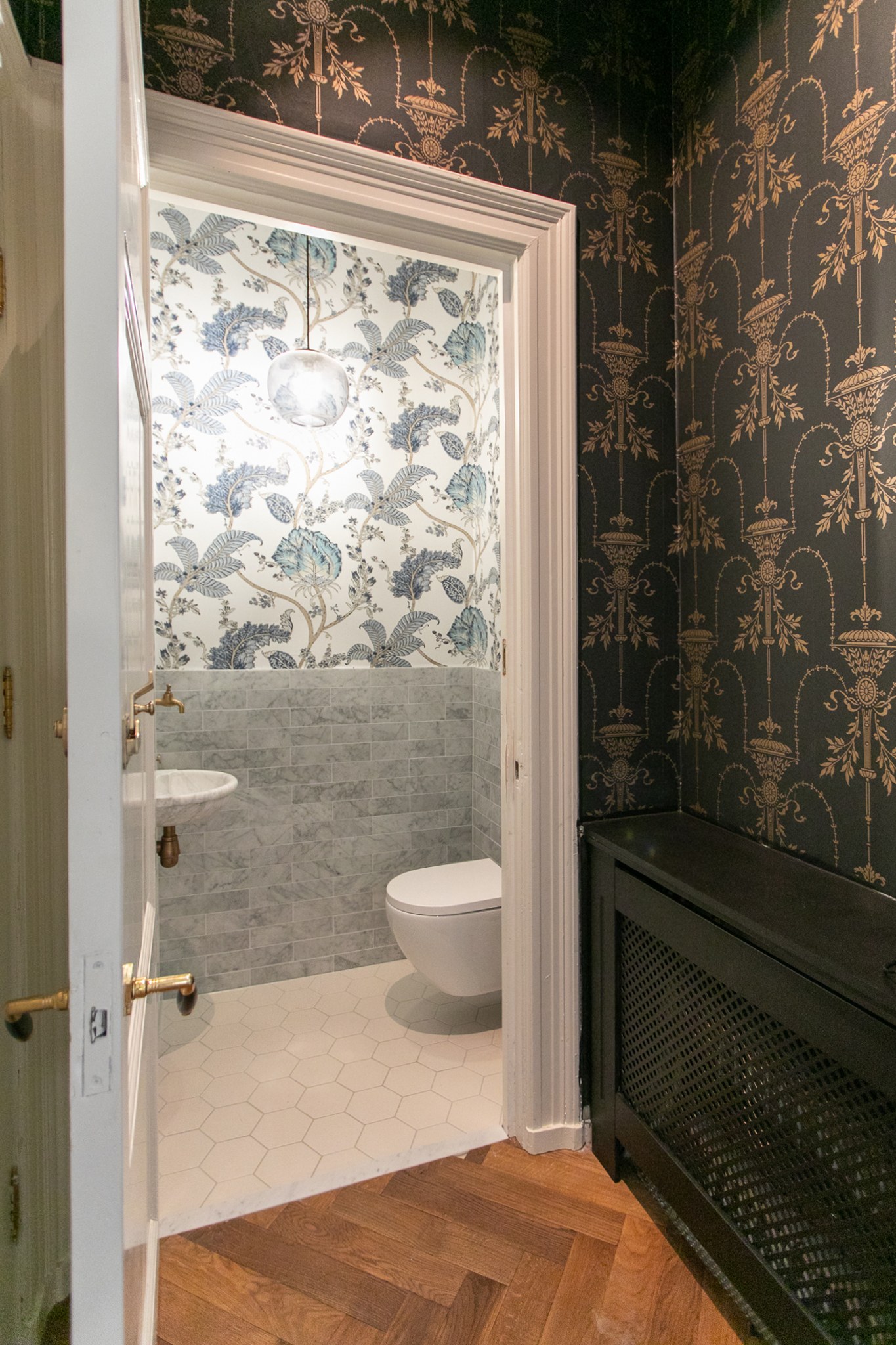 Sfeervolle doorgang naar een toilet met donker botanisch behang, zwarte radiatorombouw, marmeren lambrisering en warme visgraatvloer.