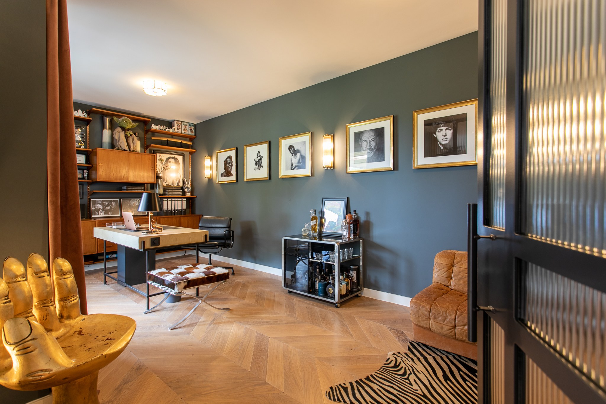 Sfeervolle werkkamer met donker blauwgrijze wand, houten bureau en boekenkast, zwart stalen ribbelglaswand, visgraatvloer en goudkleurige designstoel.