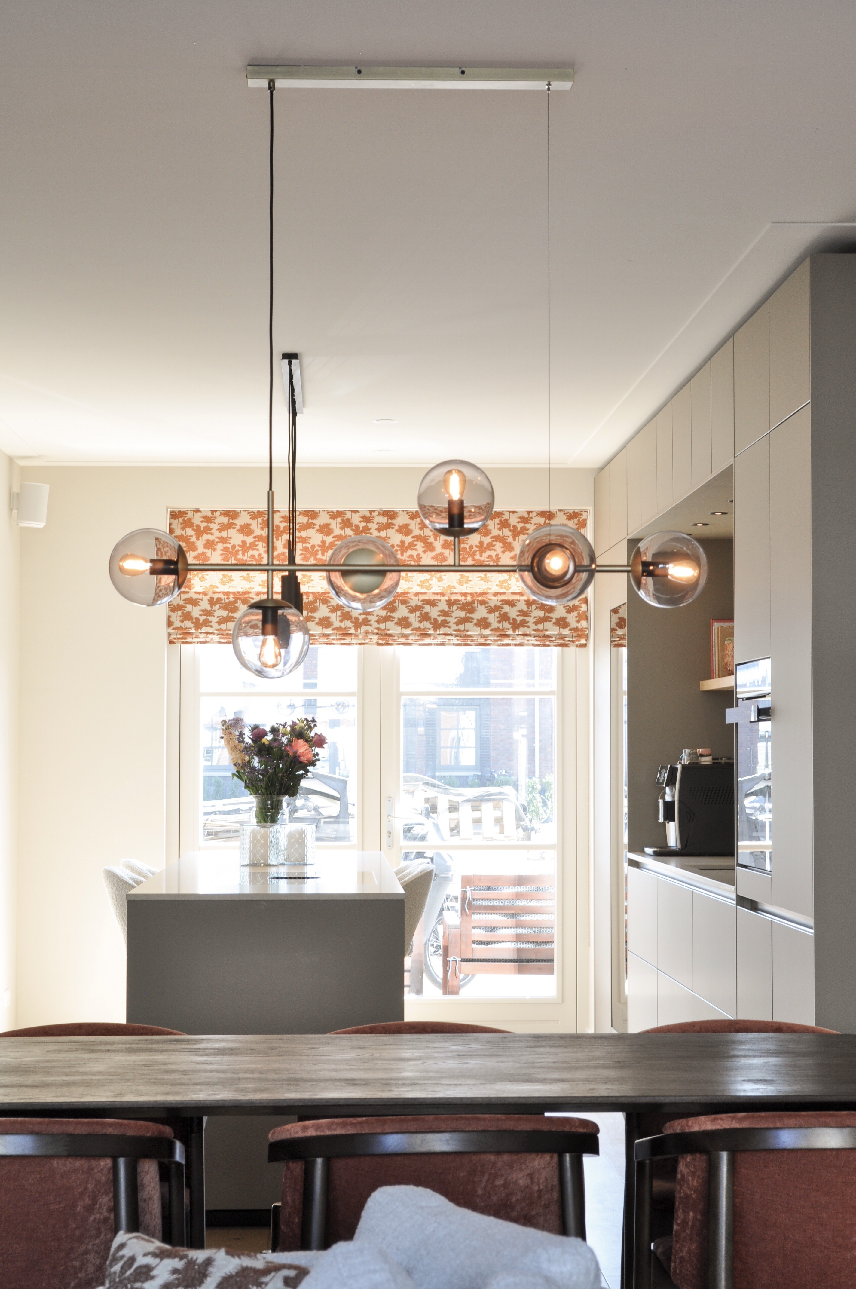 Zicht vanuit de eetkamer met glazen bolhanglampen naar een taupe maatwerkkeuken.