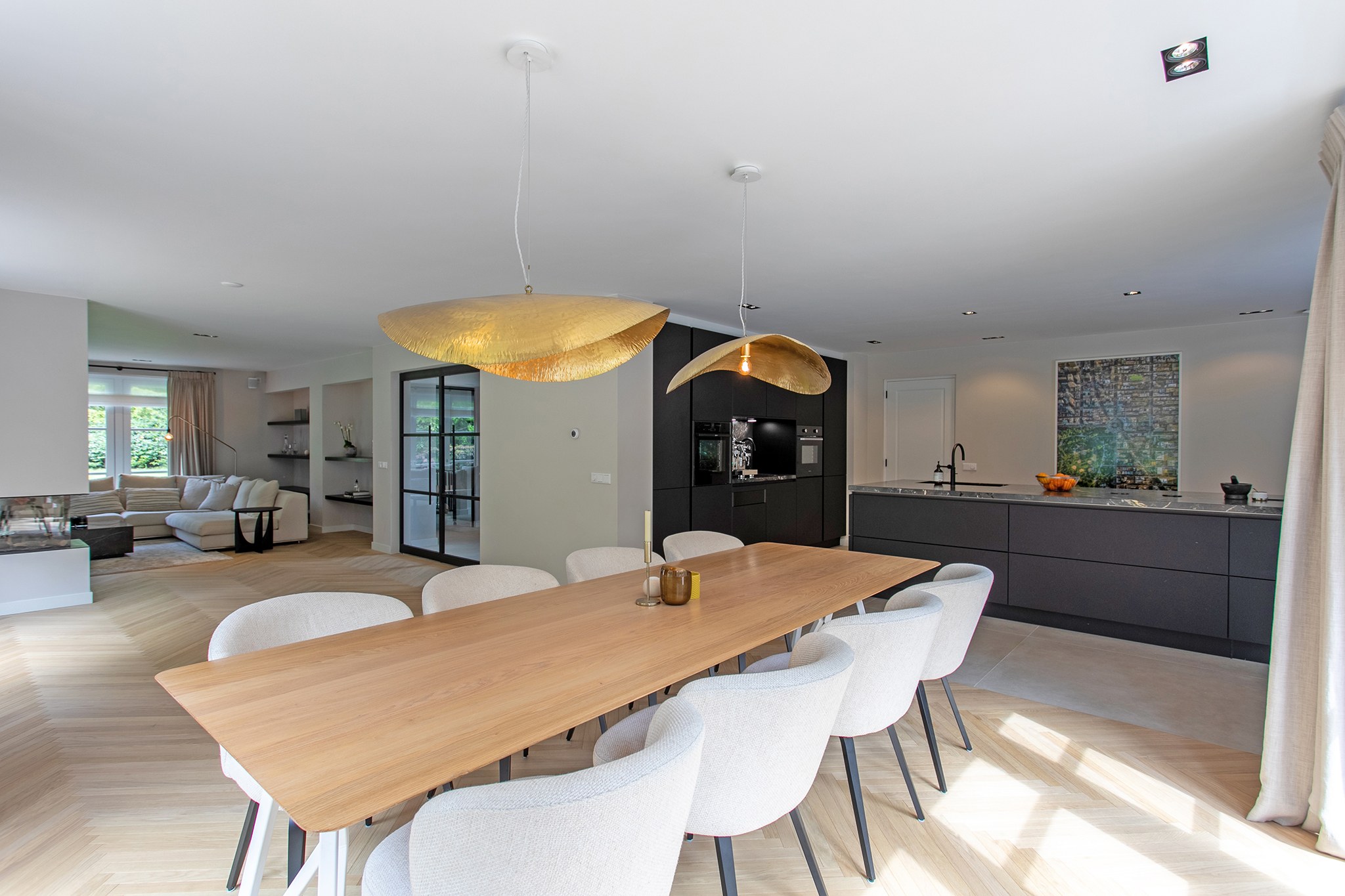 Lichte eethoek in Blaricum met grote houten tafel, crèmekleurige stoelen, organische gouden hanglampen, matzwarte keuken en neutraal grijsbeige palet.