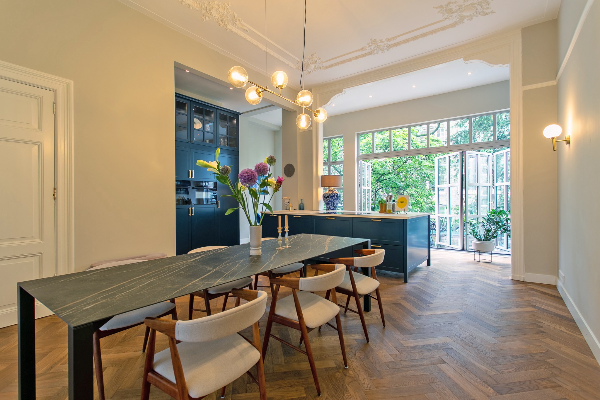 Warm moderne eetkamer met donker natuurstenen tafel, lichte stoelen, visgraatvloer, donkerblauwe keuken, messing verlichting en klassieke plafondornamenten.