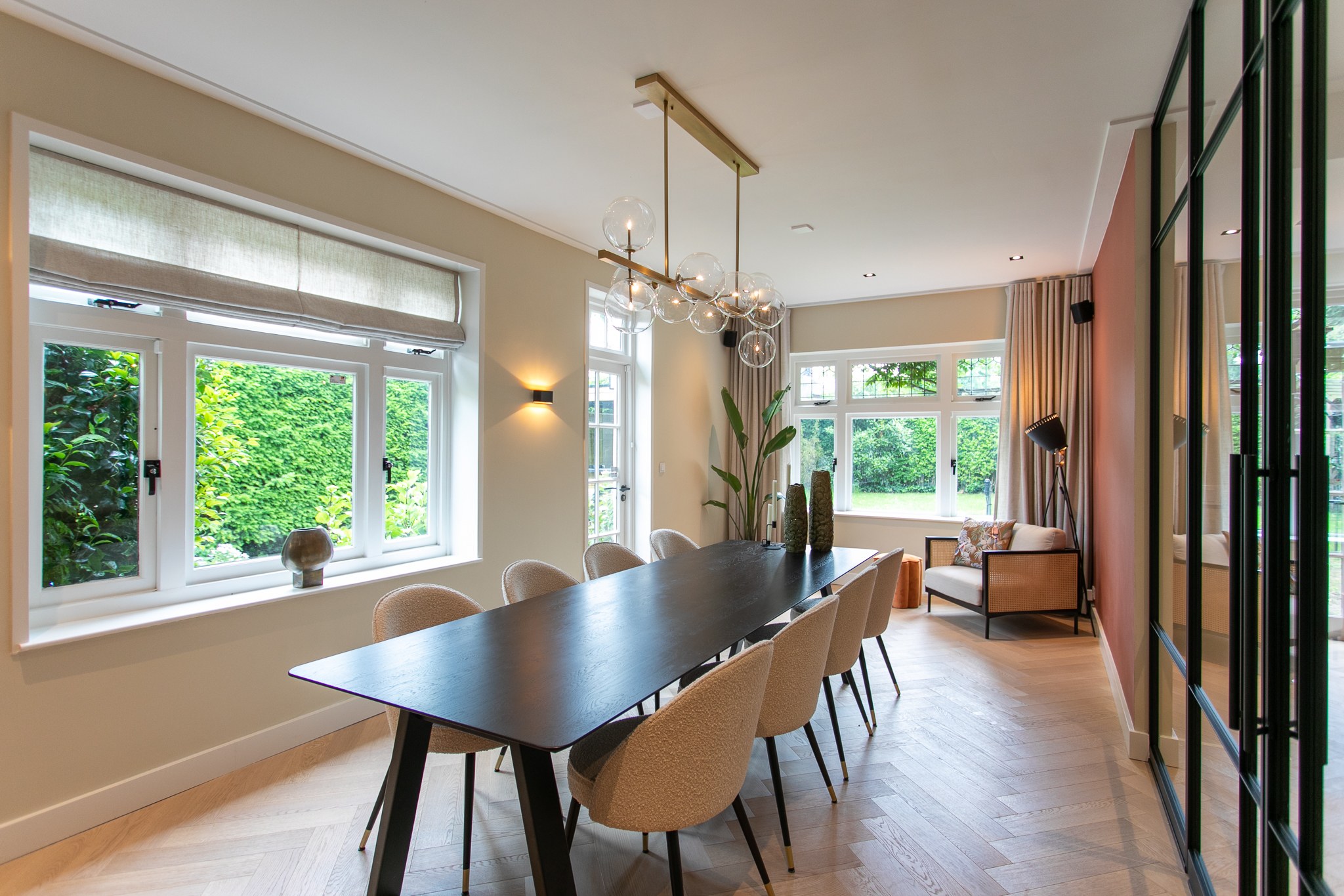 Lichte eetkamer met lange zwarte tafel, bouclé stoelen, visgraatvloer, terracotta accentwand, glazen bolverlichting en zwarte stalen scheidingswand.
