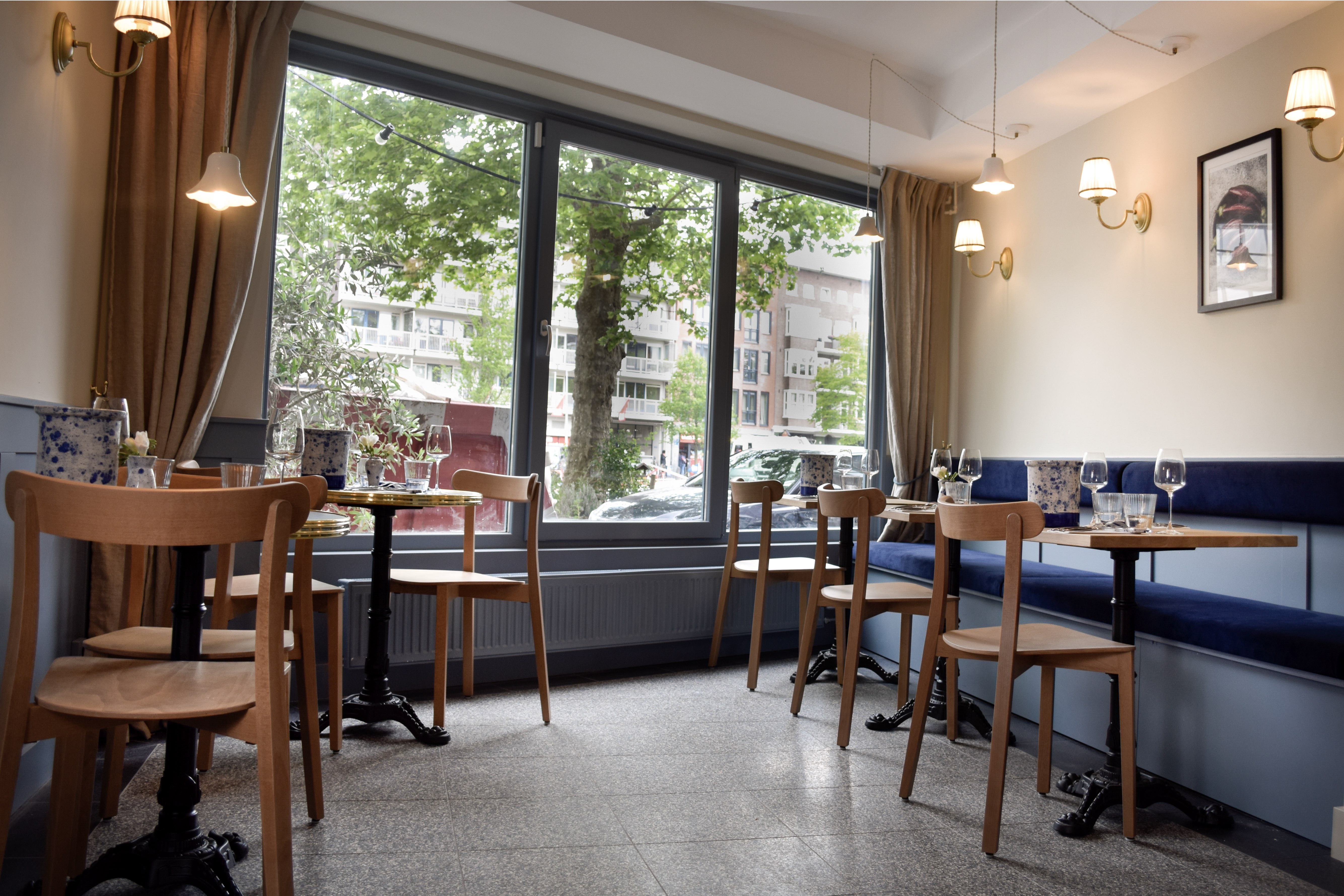 Lichte eetzaal met blauwe wandbank, houten stoelen, ronde tafels en kamerbrede ramen.