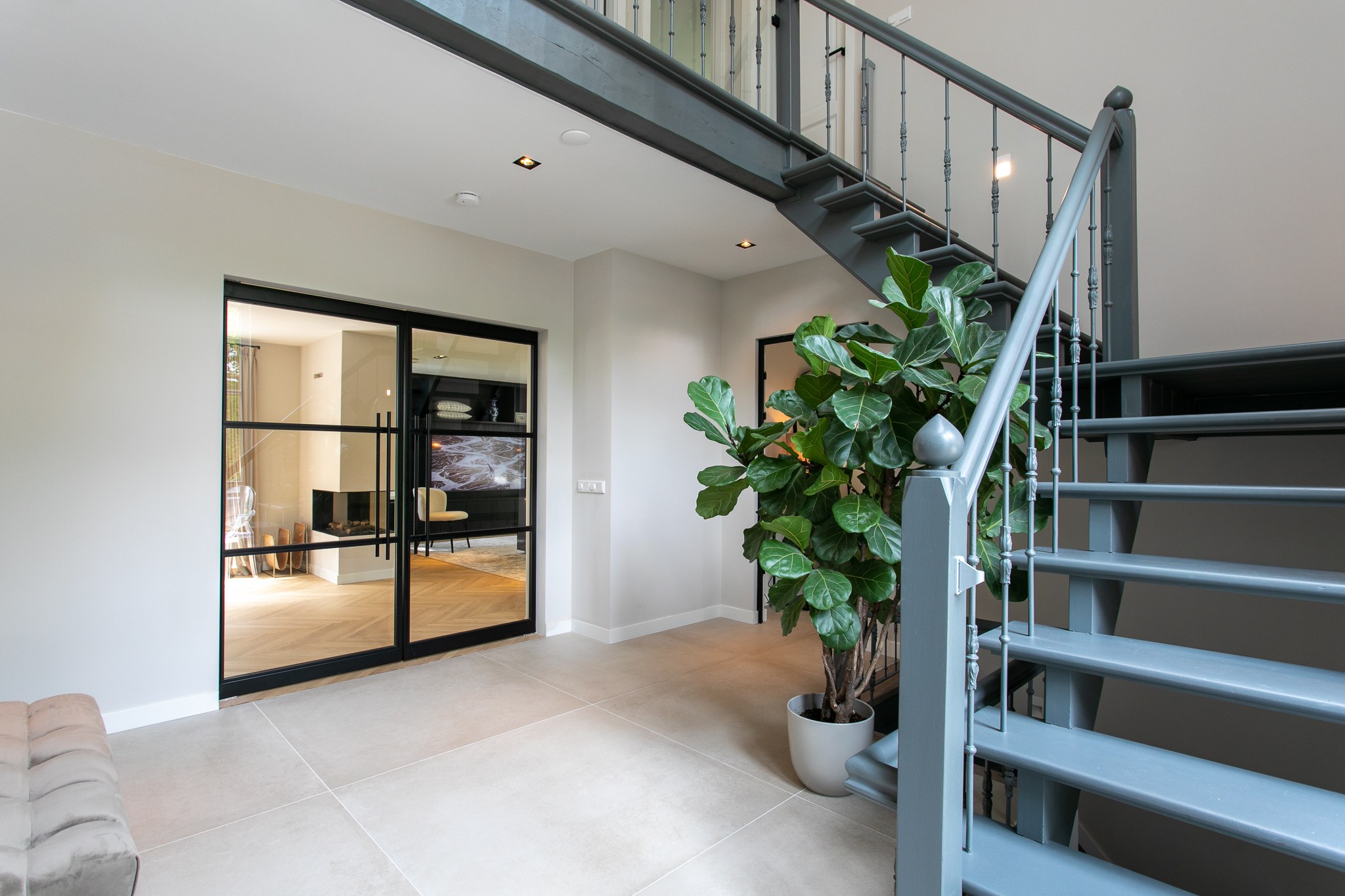 Moderne entree met open zwart stalen trap, glazen balustrade, zwarte stalen taatsdeuren, grote groene plant en lichte grootformaat tegels.