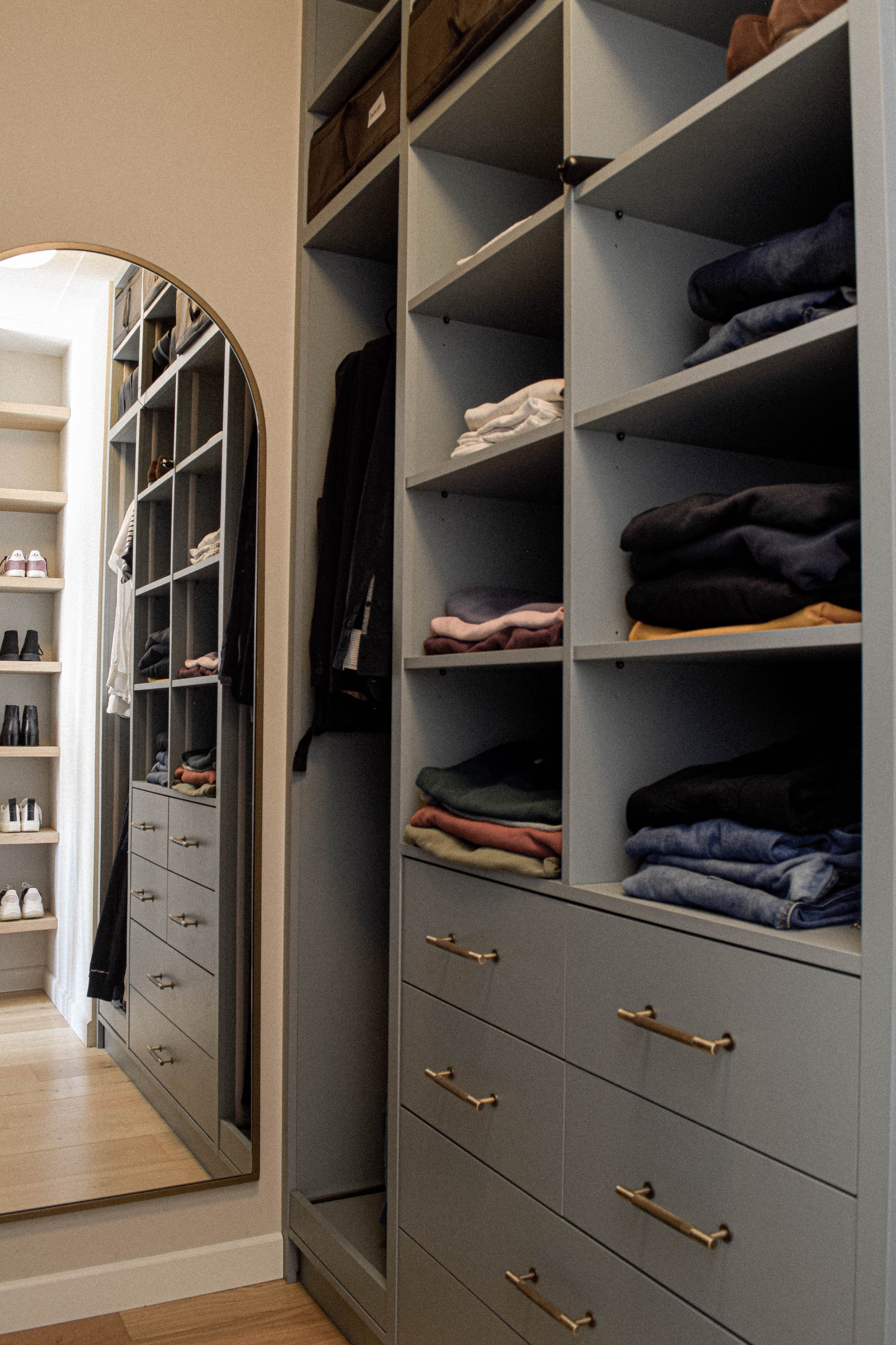 Open grijsgroene maatwerkgarderobe met messing grepen en ovale spiegel.