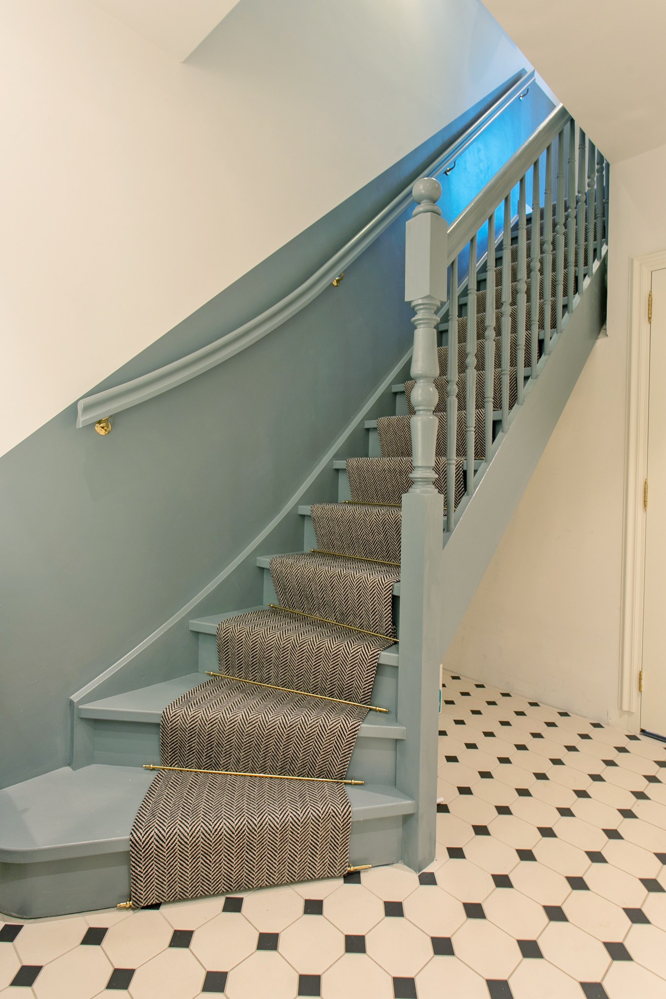 Klassieke trap met blauwgrijze houten balustrade en lambrisering, grijs geweven traploper met messing roedes en zwart-witte tegelvloer.