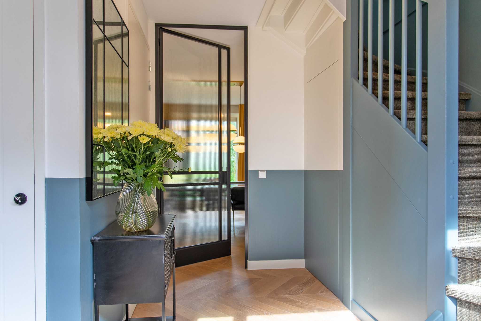 Moderne hal met blauwgrijze hoge lambrisering, witte wanden, visgraatvloer, zwarte stalen glazen deur en vintage console met bloemen.