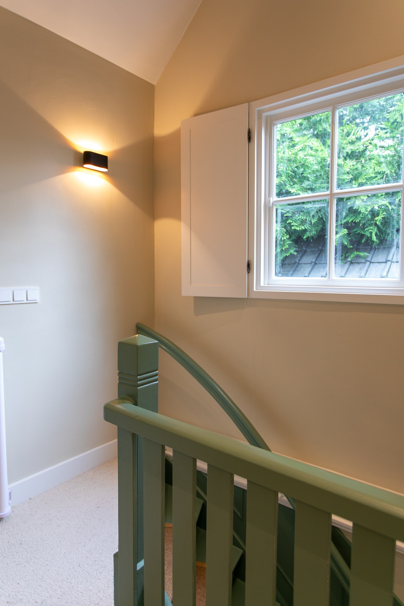 Lichte trapvide met grijsgroene houten balustrade, warme greige wanden, zacht tapijt, wit kozijn en moderne zwarte wandlamp.