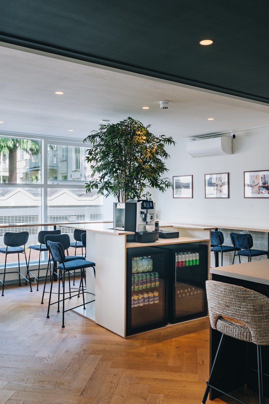 Open kantoorbar met centraal drankmeubel en koelkasten, lichte visgraatvloer, groenblauwe barkrukken, kamerhoge ramen en grote binnenboom.