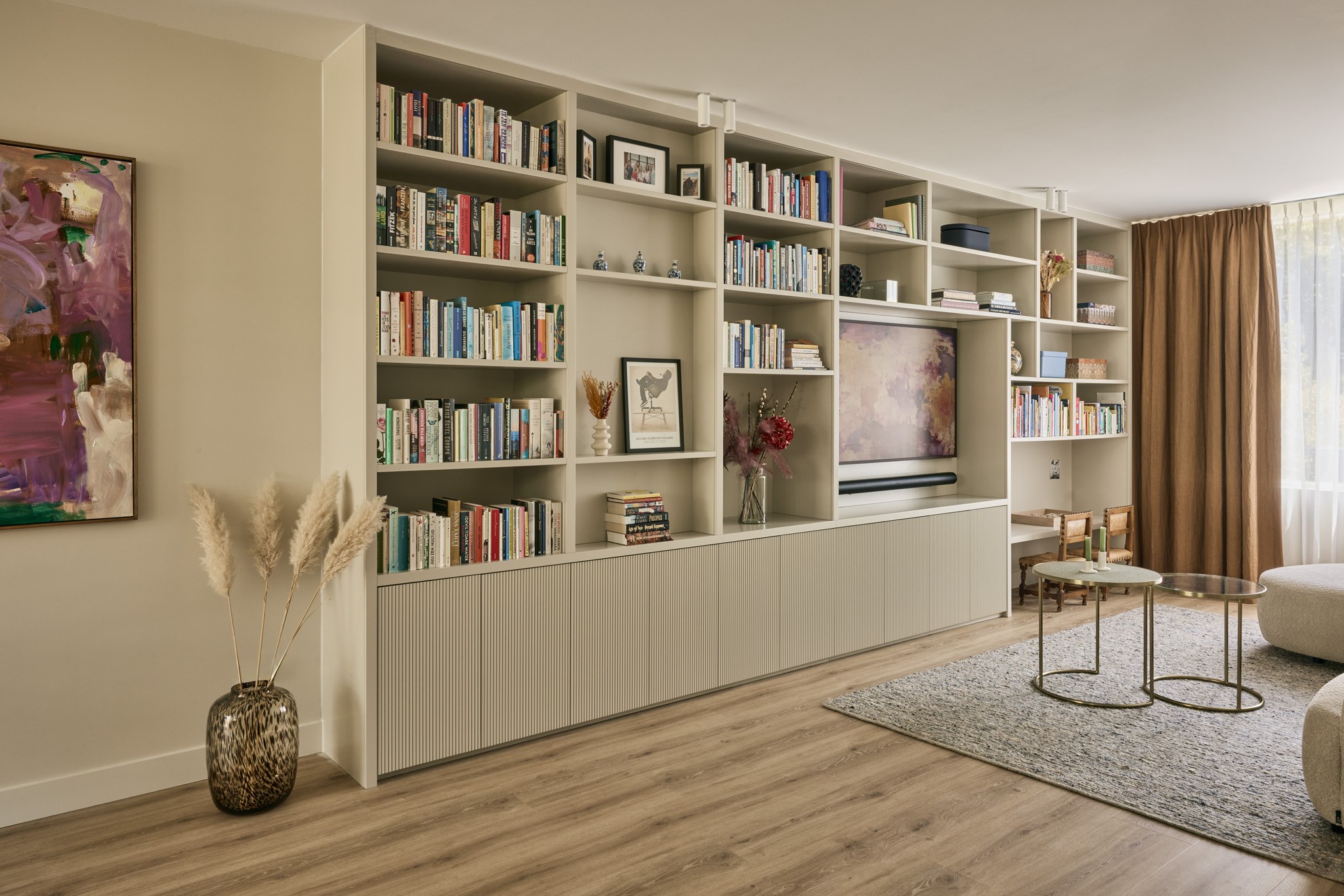 Kamerbrede beige maatwerkwand met televisie, open boekenvakken en organische crèmekleurige bank.