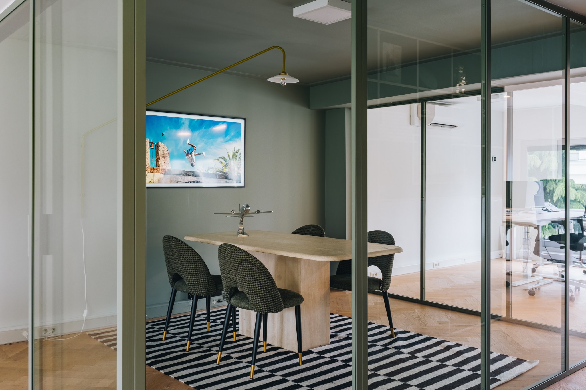 Moderne glazen vergaderruimte met groen-grijze wanden, lichte houten tafel, donkergroene stoelen, zwart-wit gestreept vloerkleed en goudkleurige wandlamp.