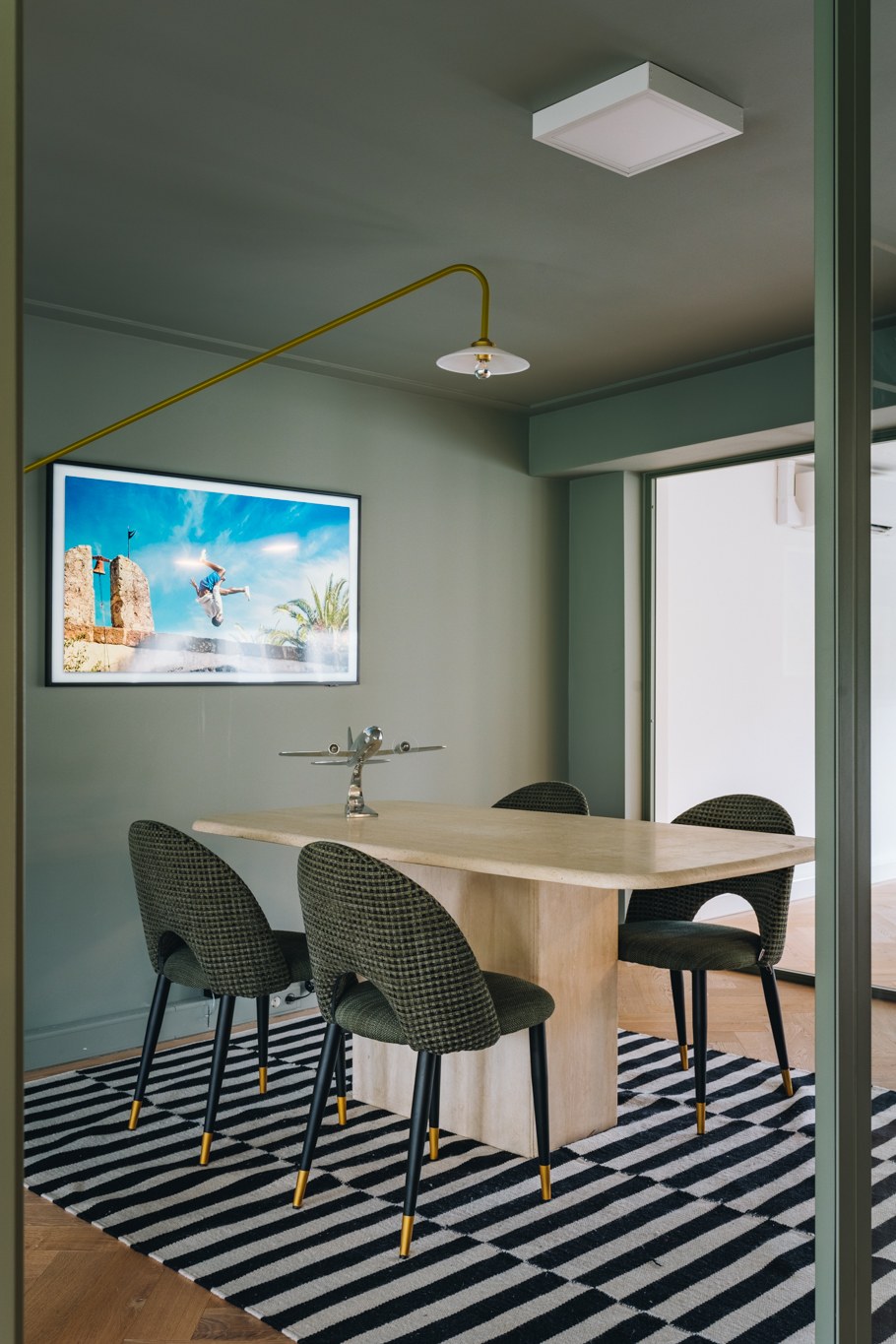 Verticale opname van glazen vergaderruimte met groen-grijze wanden en plafond, lichte houten tafel, donkergroene stoelen en zwart-wit gestreept vloerkleed.