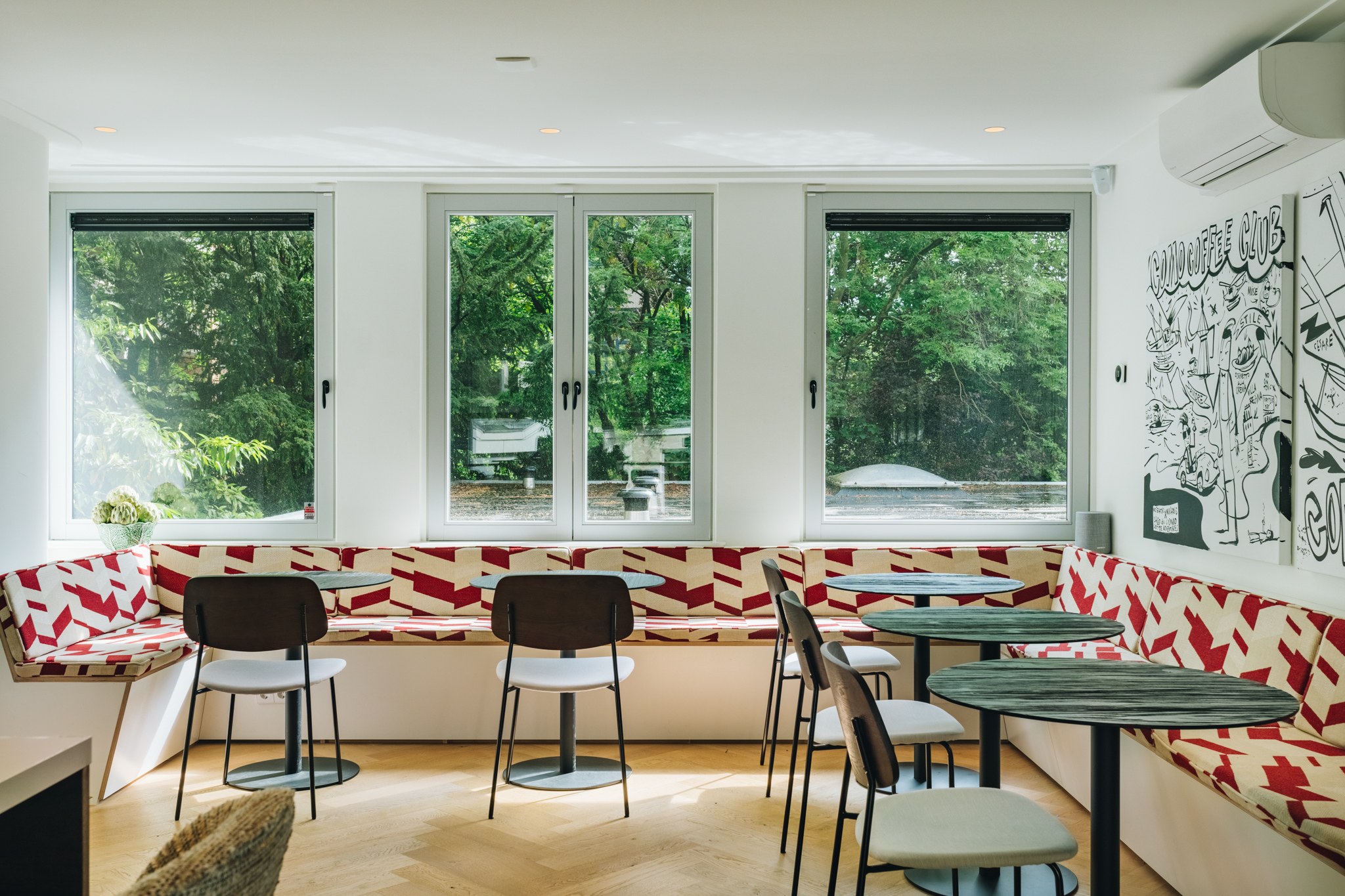 Lichte bedrijfskantine met lange ingebouwde bank in rood-wit grafische stof, kleine groene tafels, zwarte stoelen, visgraatvloer en wandillustratie.