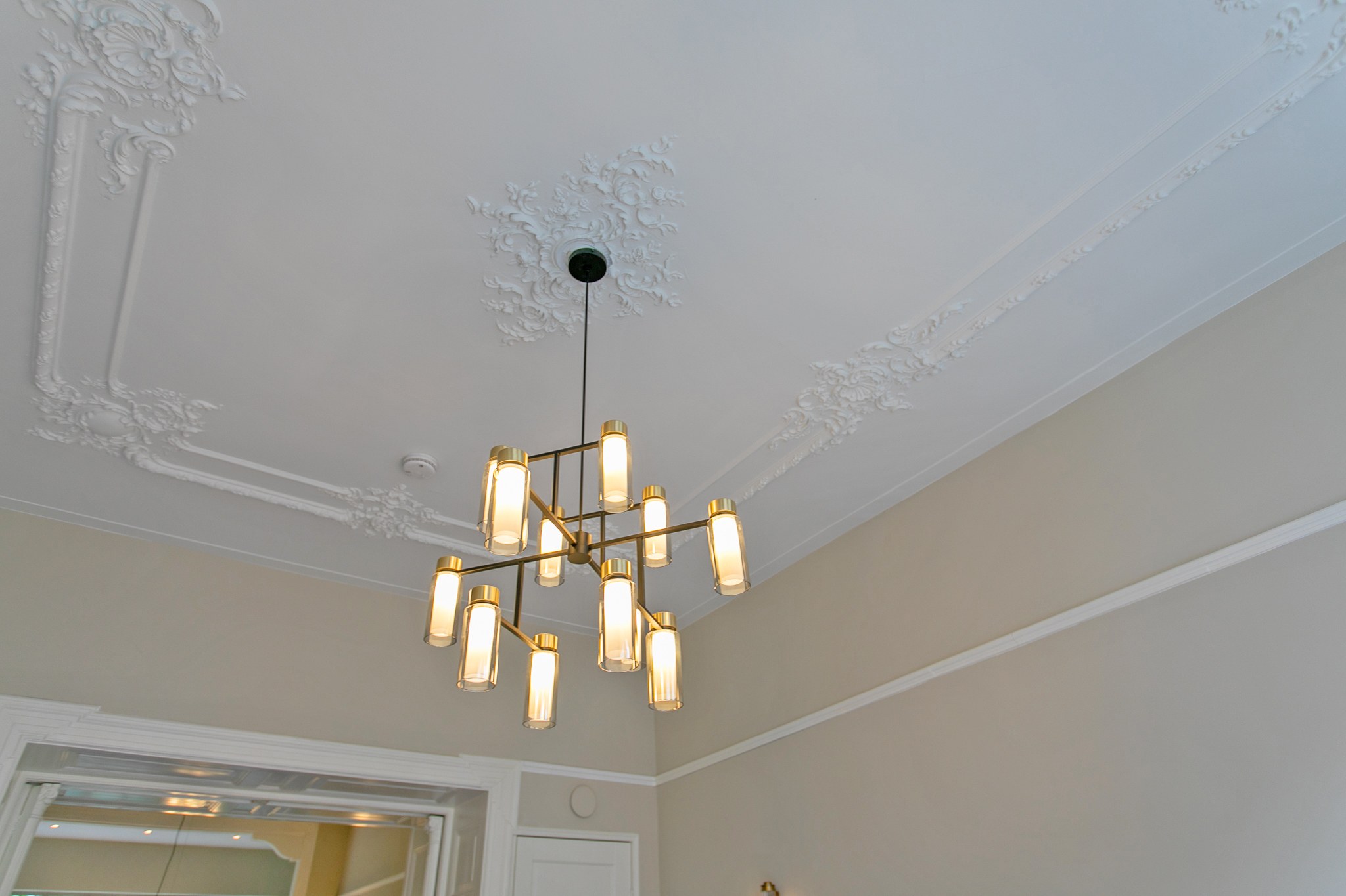Detail van klassiek wit stucwerk en plafondornamenten met een moderne geometrische messing kroonluchter tegen zacht beige wanden.