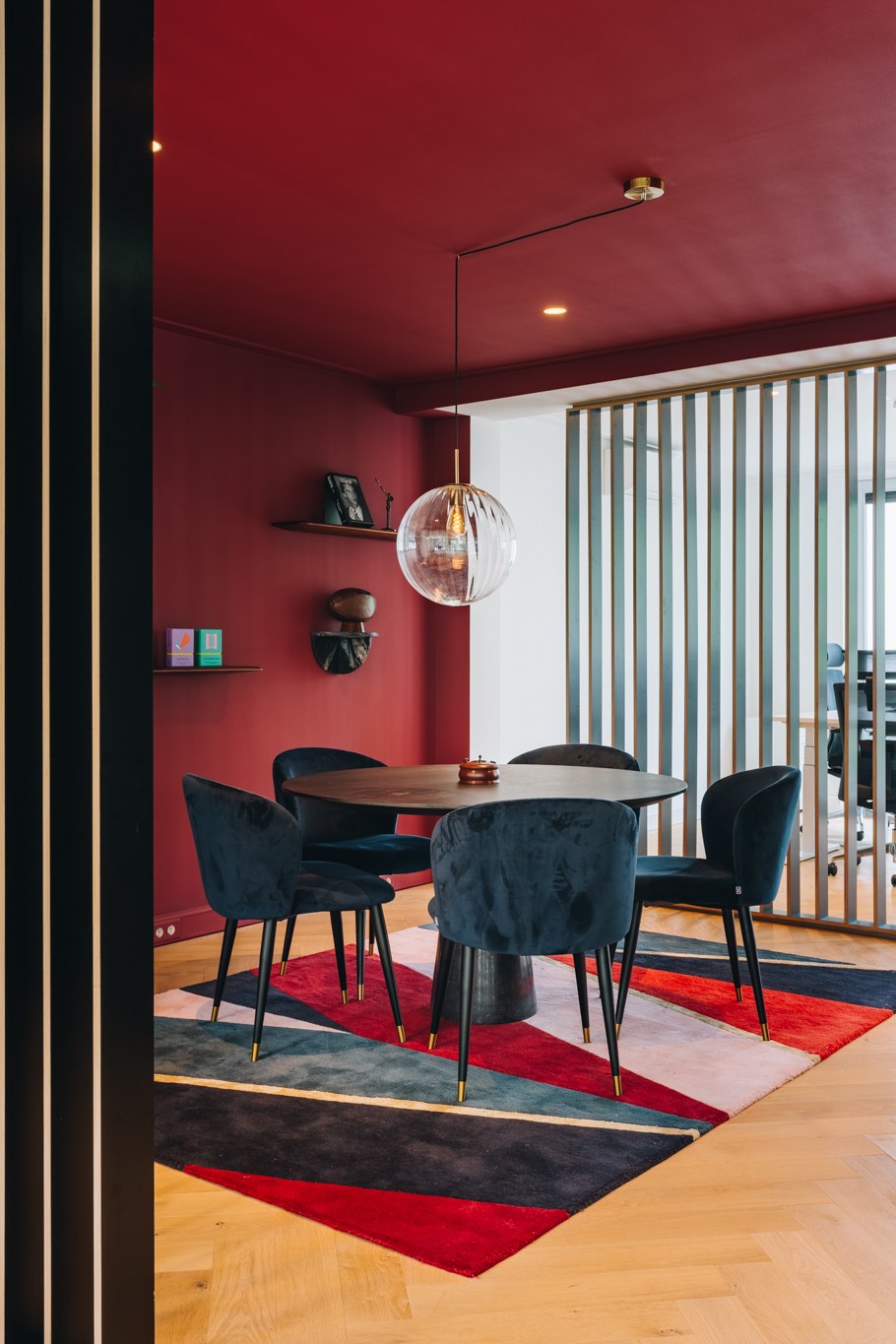 Compacte vergaderplek met diepe bordeauxrode wanden, zwarte ronde tafel, blauwe fluwelen stoelen, glazen bolhanglamp en zwarte verticale lamellen.