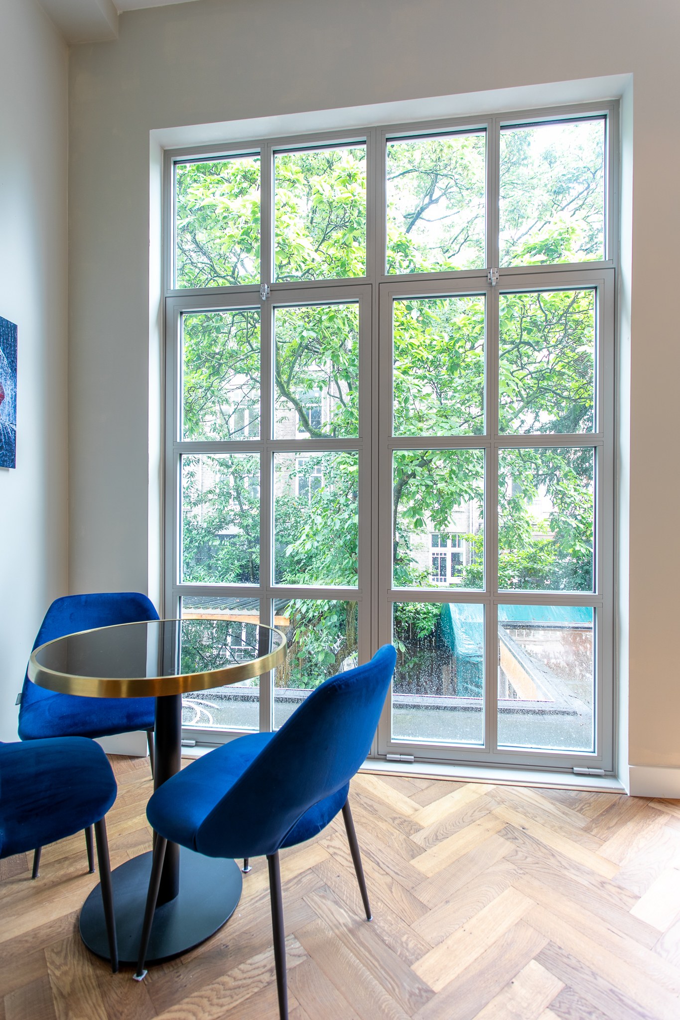Elegante zithoek met hoge klassieke raampartij, twee kobaltblauwe stoelen, ronde marmeren tafel en warme visgraatvloer.