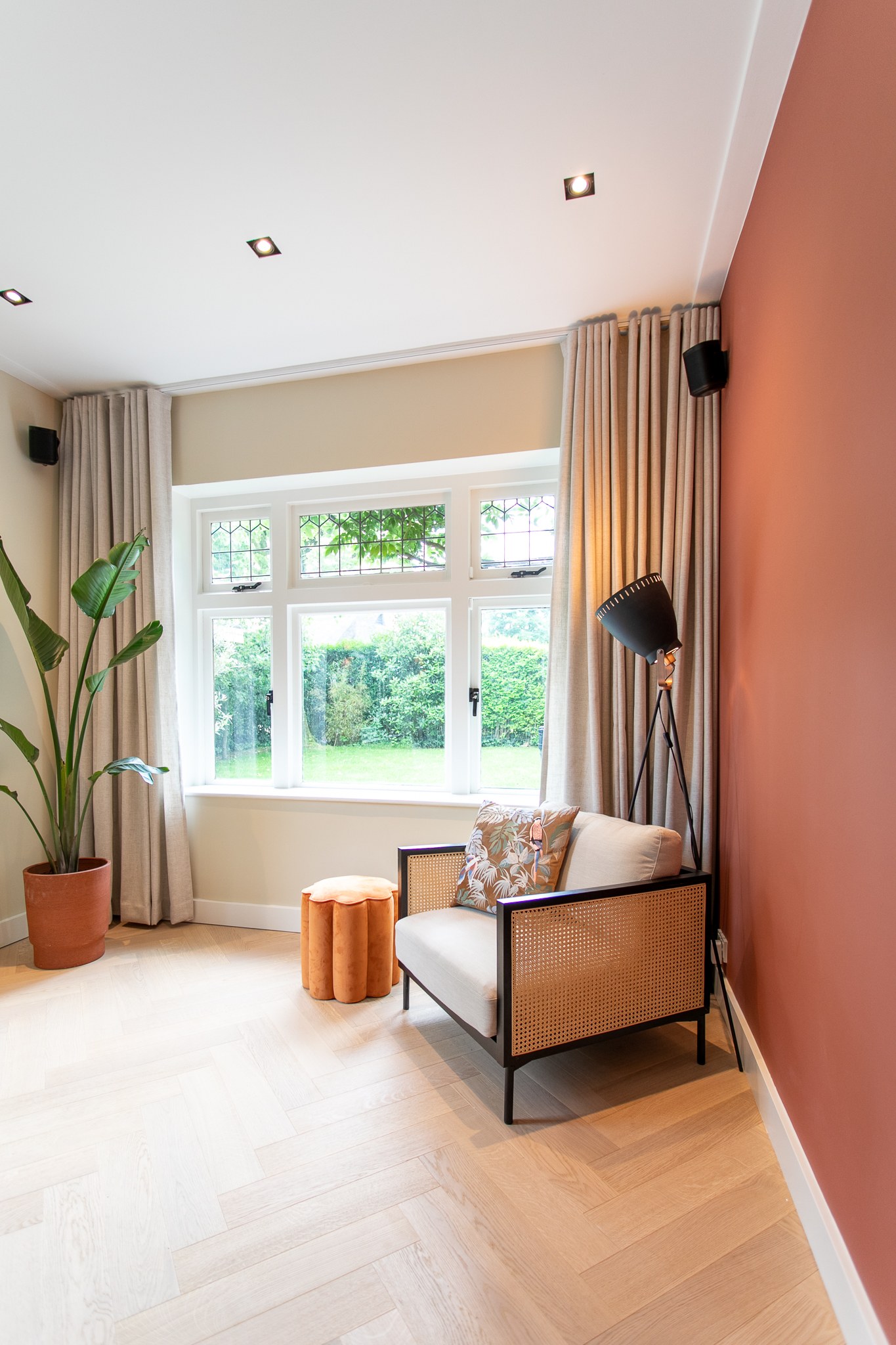 Rustige zithoek met terracotta wand, cognackleurige fauteuil, sculpturale houten bijzettafels, kamerhoge beige gordijnen en grote groene plant.