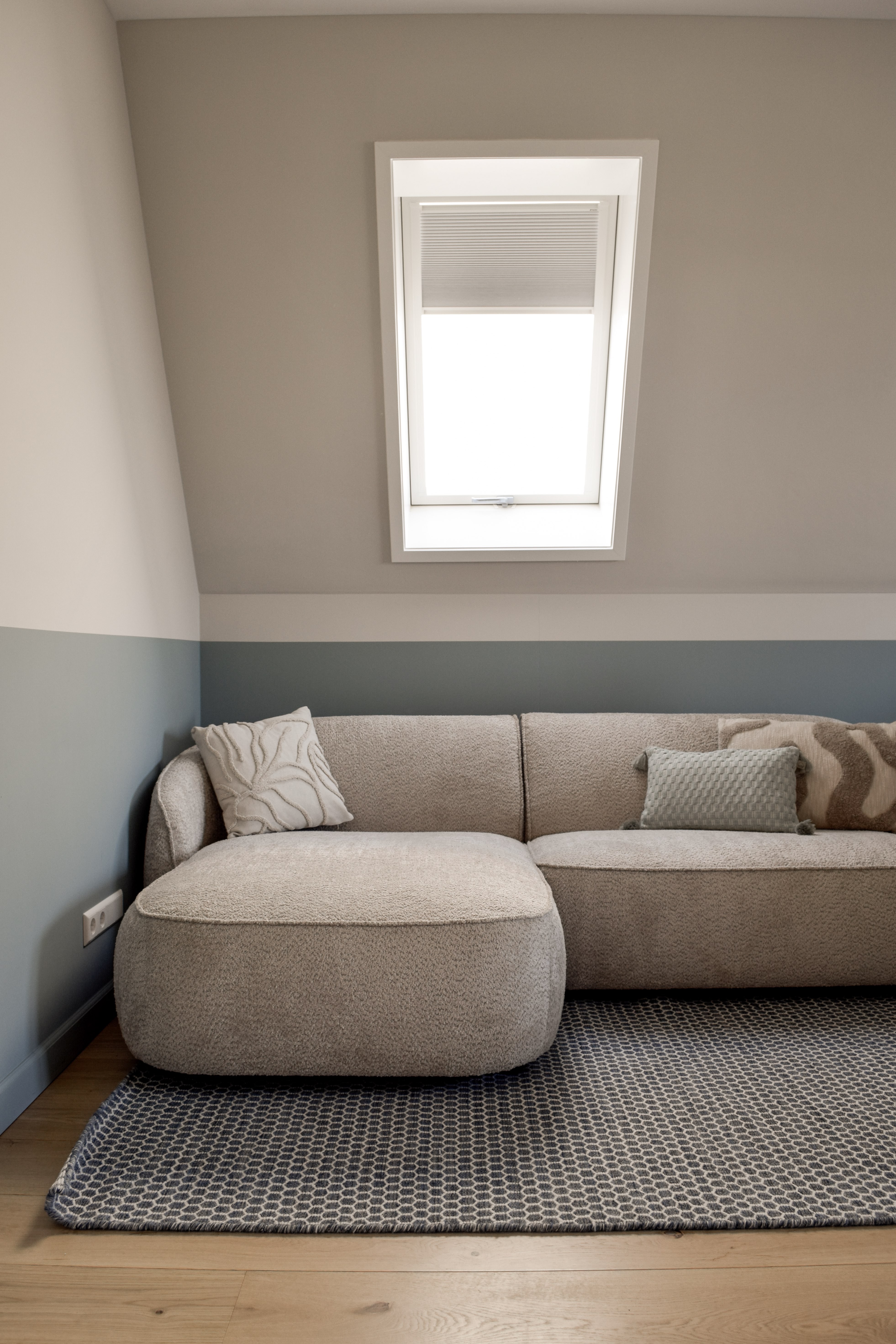 Zolderkamer met blauwgrijze wanden, taupe hoekbank, dakraam en zachte neutrale materialen.
