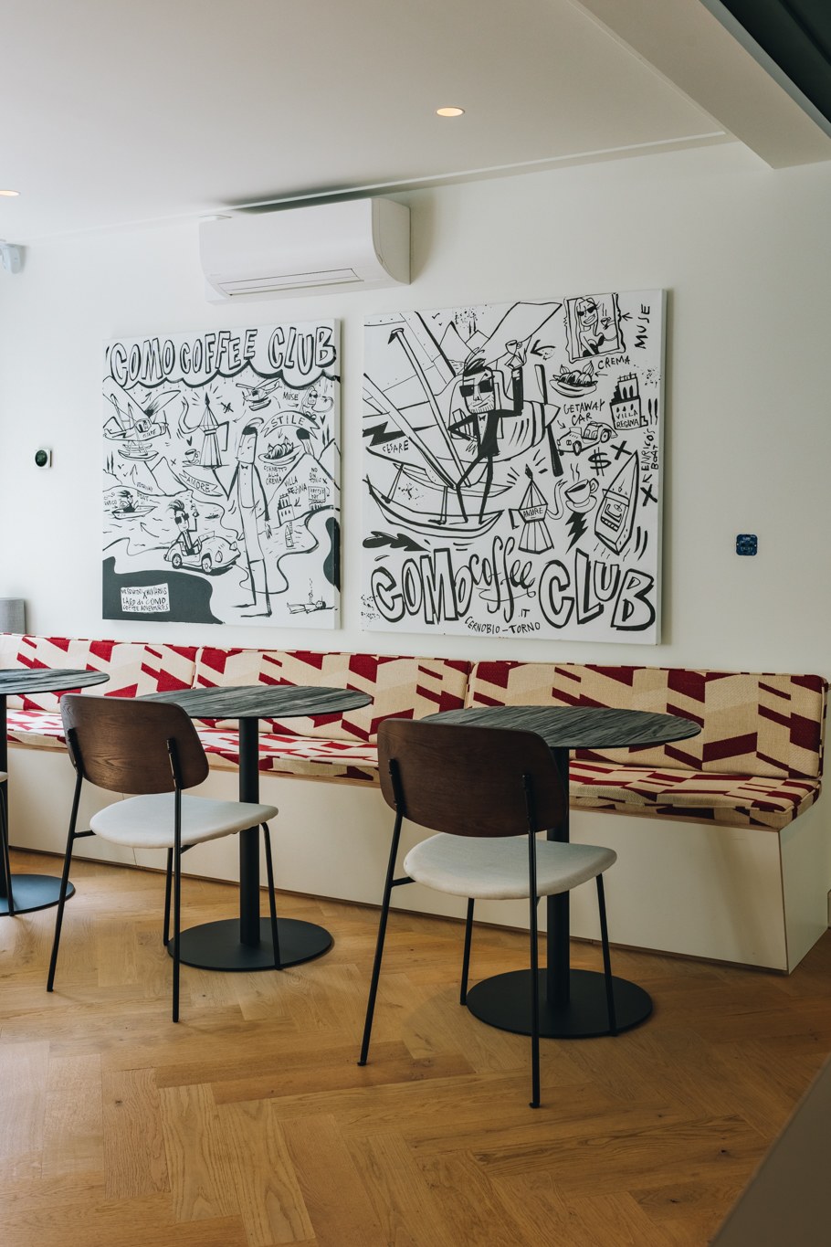 Compacte eetnis in kantoor met rood-wit gestoffeerde maatwerkbank, kleine ronde tafels, zwarte stoelen, grafische wandkunst en visgraatvloer.