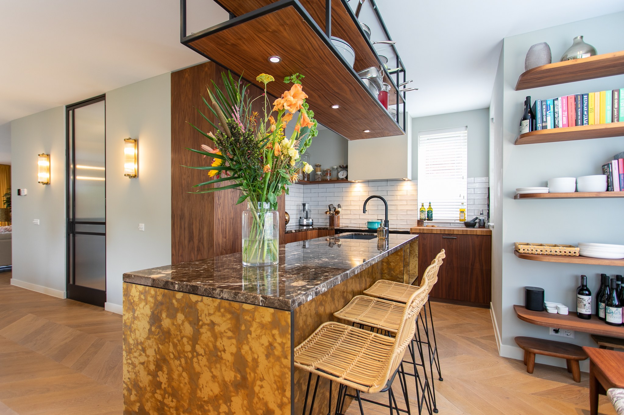 Eclectische keuken met uitgesproken bruin natuurstenen eiland, walnoot plafondrek en kasten, rotan barkrukken, zwarte kraan en open houten wandplanken.