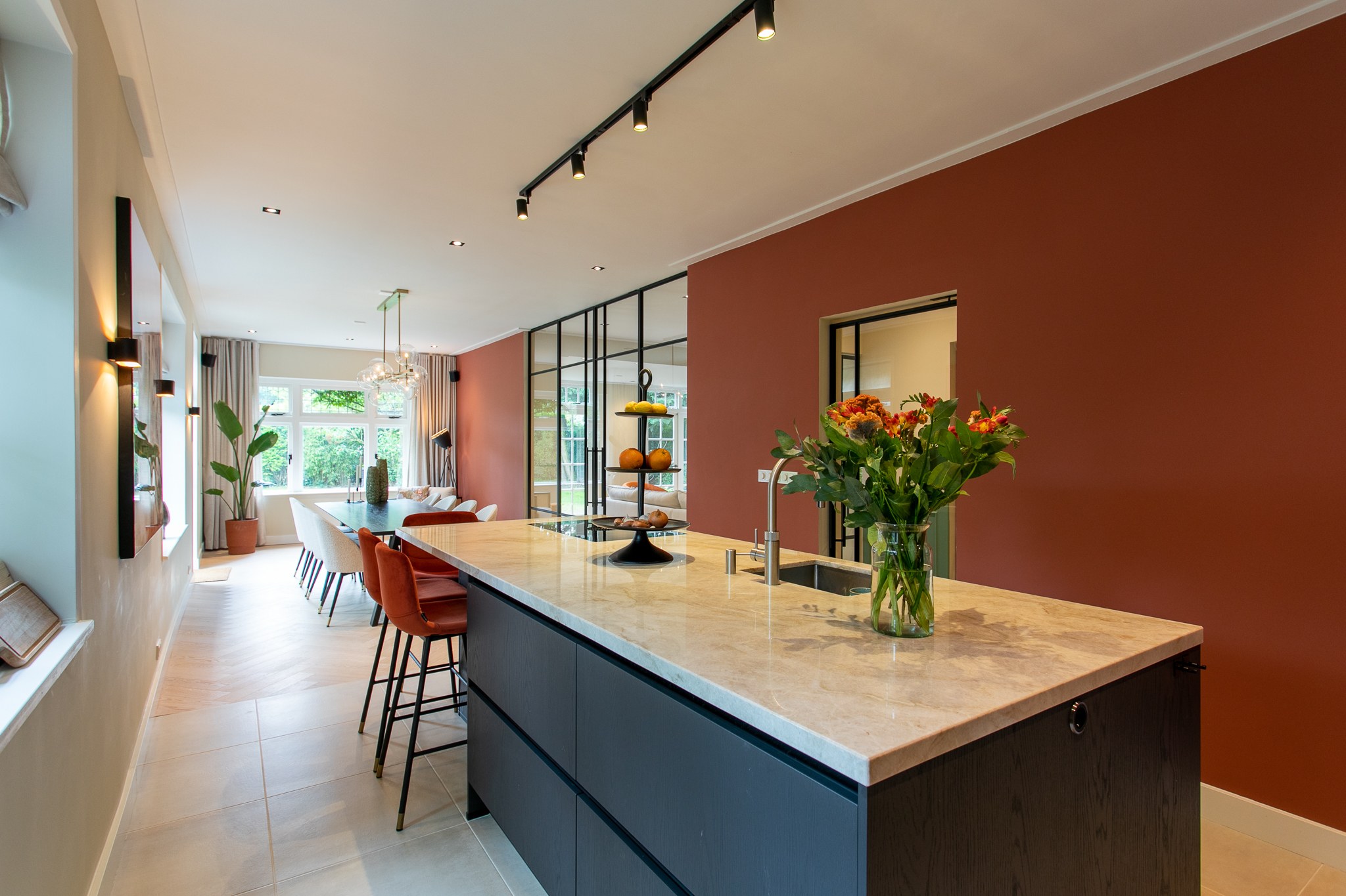 Warme leefkeuken met donker kookeiland, licht natuurstenen blad, diepe terracotta accentwand, zwarte stalen glazen scheidingswand en lichte vloer.