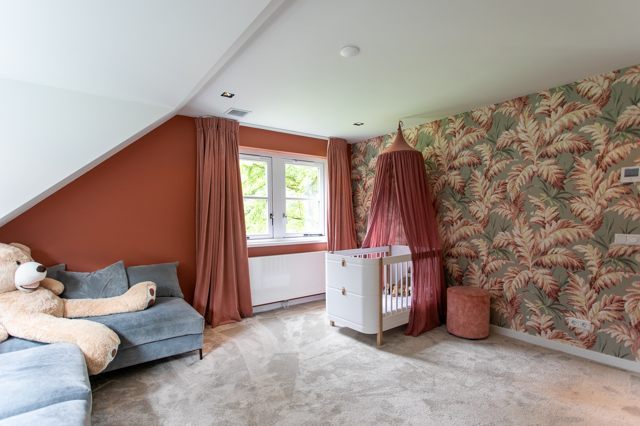 Warme kinderkamer met terracotta wand en gordijnen, botanisch behang, wit ledikant met oudroze hemel, grijsblauwe bank en zacht vloerkleed.