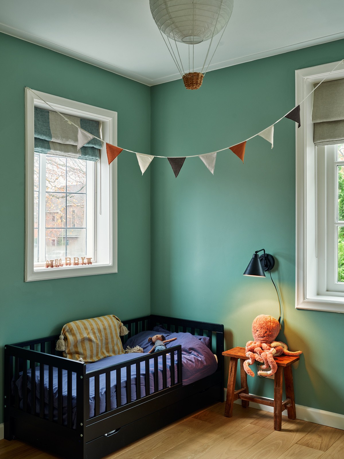 Knusse kinderkamer in Bussum met zeegroene wanden, donker houten bed, zachte grijze vouwgordijnen en warme oker- en oranjetinten.