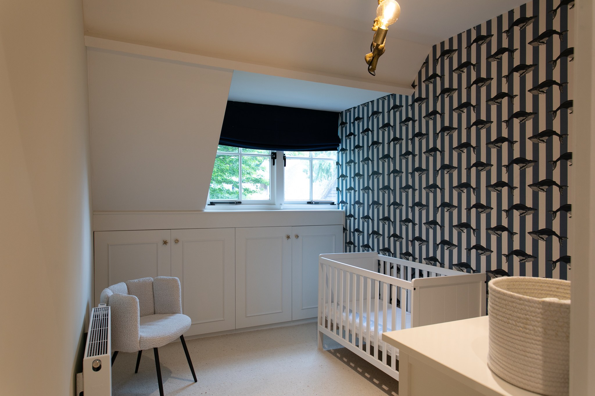 Kinderkamer onder schuin dak met zwart-wit grafisch behang, wit ledikant, witte maatwerkkasten, licht tapijt en zwarte vouwgordijnen.