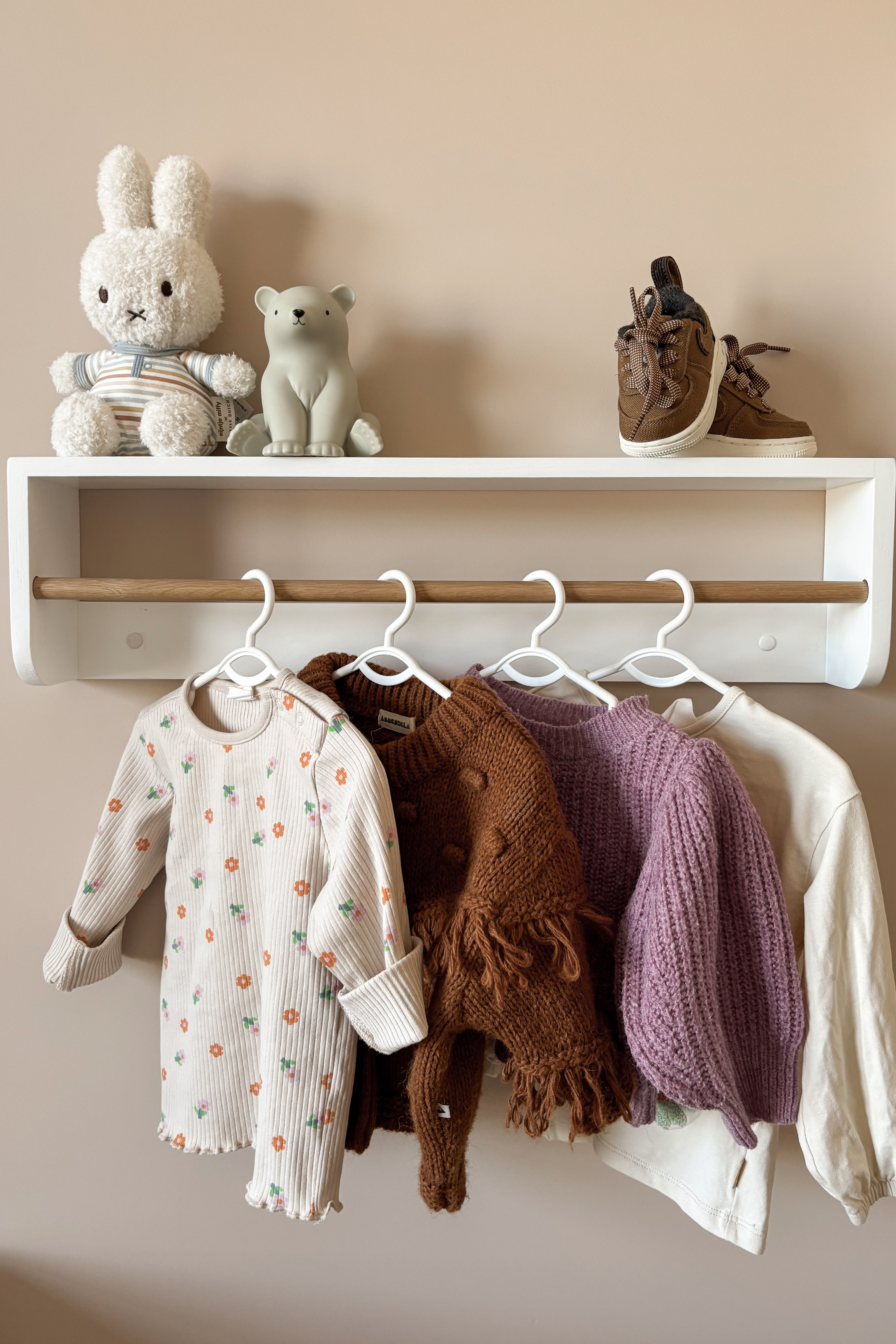 Detail van kinderkleding aan een rek in een babykamer met beige, bruin en lila tinten.