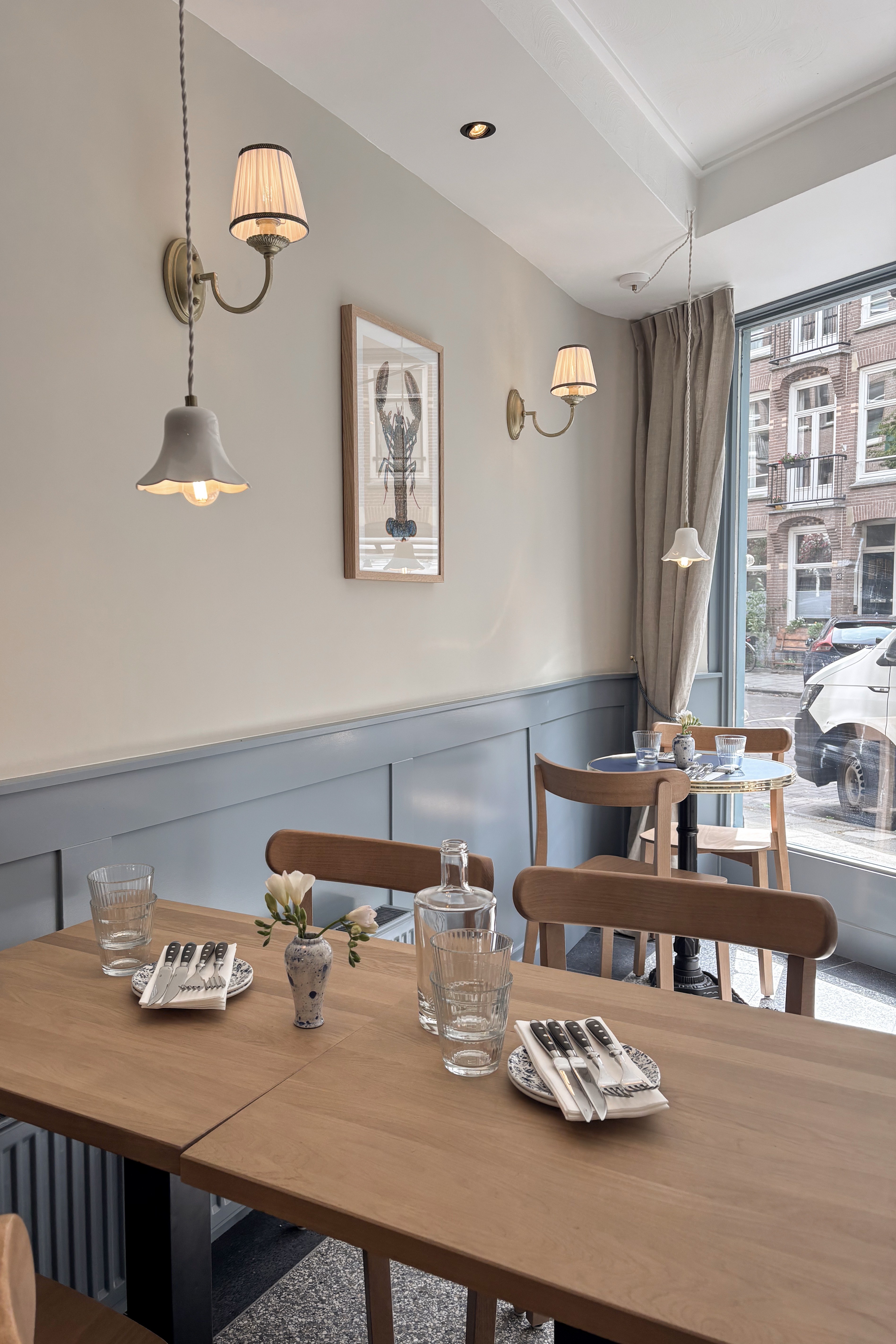 Gedekte lange houten tafel met houten stoelen, blauwgrijze lambrisering en klassieke wandlampen.