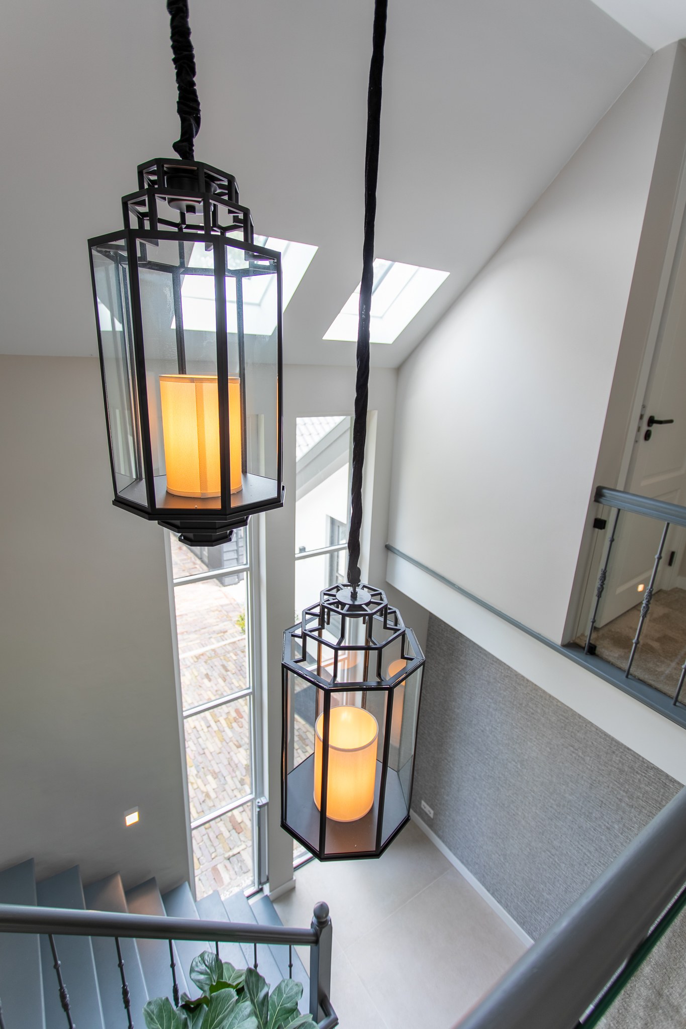 Detail van twee grote zwarte glazen lantaarnlampen in een lichte vide met open trap, glazen balustrade en dakramen.