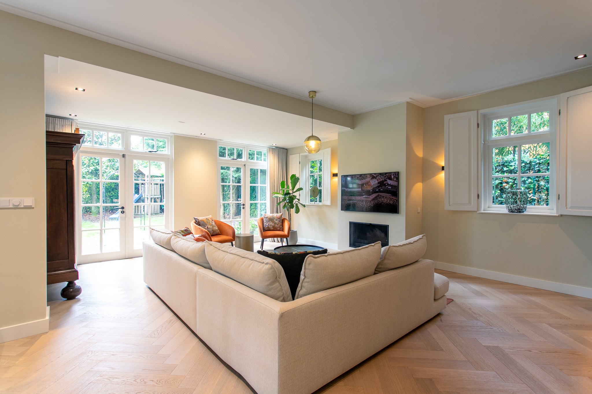 Open woonkamer met lichte hoekbank, visgraatvloer, ingebouwde haard, greige wanden, cognackleurige fauteuil en grote klassieke ramen naar de tuin.