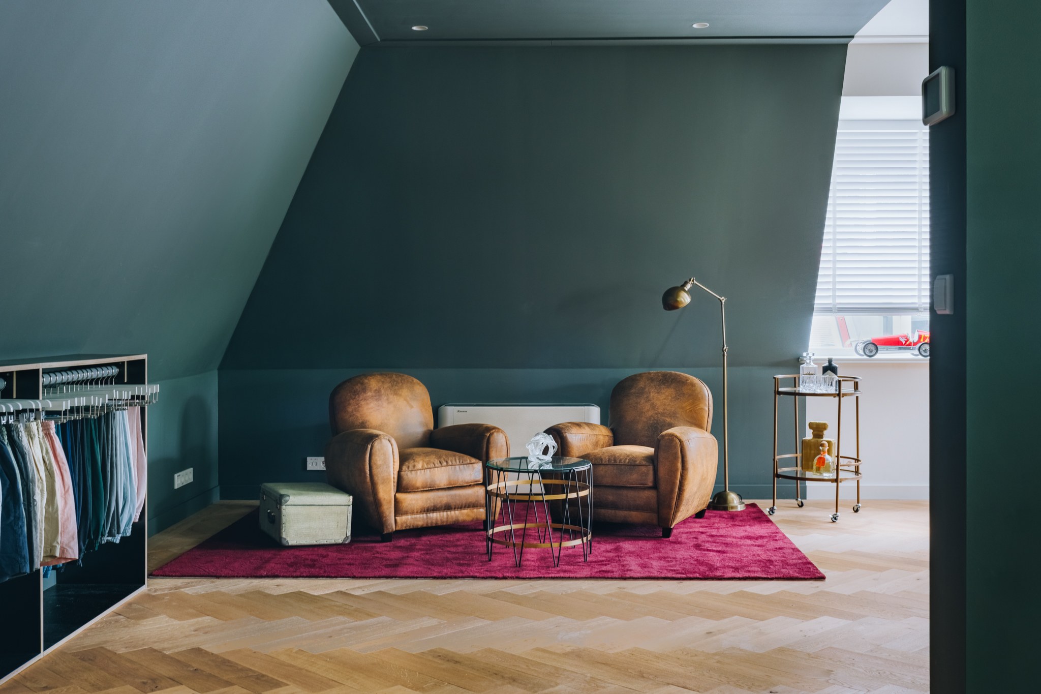 Sfeervolle lounge onder schuin dak met donker petrolgroene wanden, twee cognackleurige leren fauteuils, magenta vloerkleed, visgraatvloer en open kledingrek.