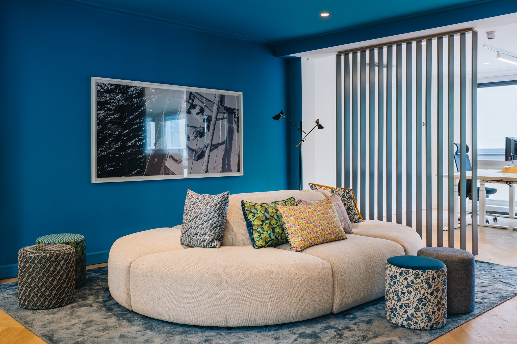 Kleurrijke kantoorlounge met diepe petrolblauwe wanden en plafond, organische crèmekleurige bank, patroonpoefs, blauw vloerkleed en houten lamellenscheiding.
